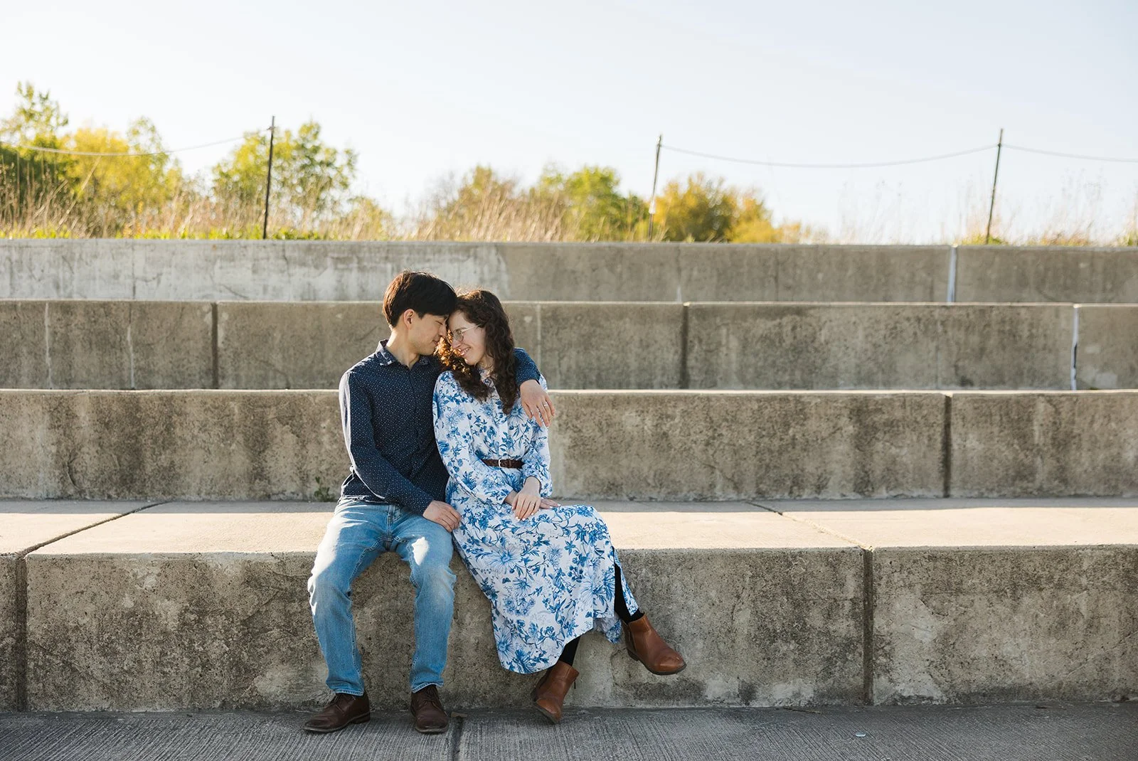 A young couple sitting on concrete steps outdoors, leaning close and smiling at each other on a sunny day.