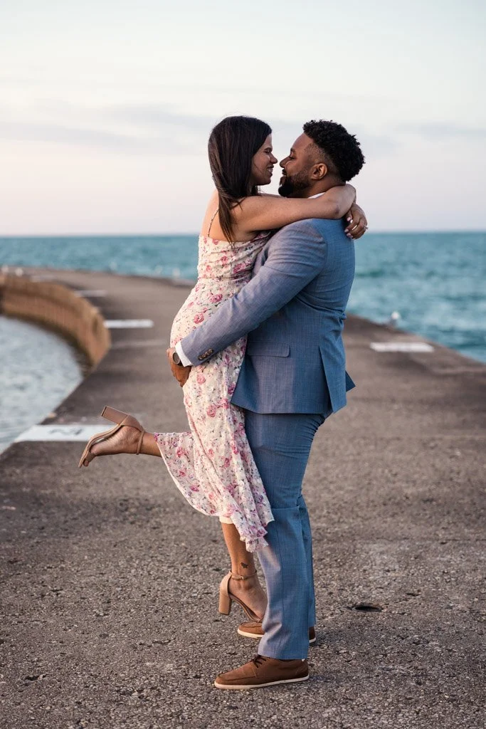 A couple embracing on a pier by the water, with the woman lifting her legs and wearing a floral dress and high heels, the man in a grey suit and brown shoes, during sunset.