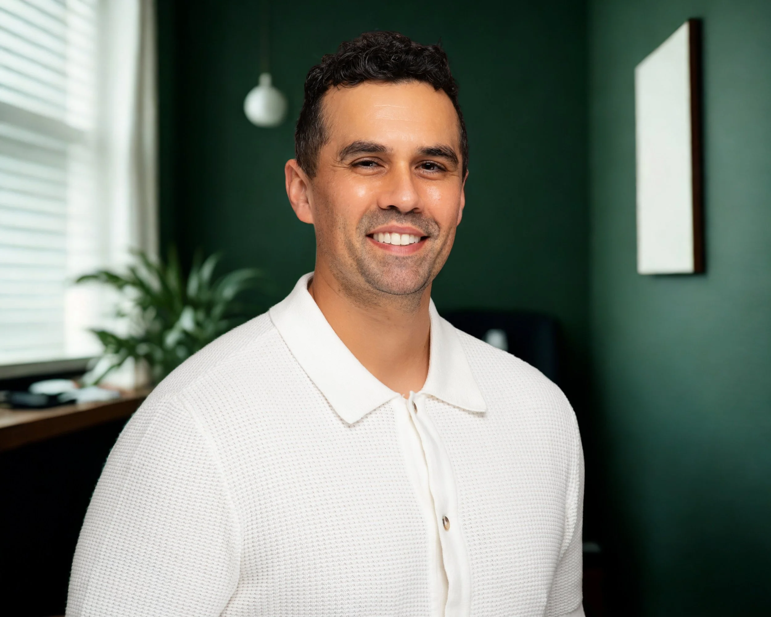 Therapist named Luis smiling with dark curly hair wearing a cream sweater standing in a modern office environment with green and white walls and plants.
