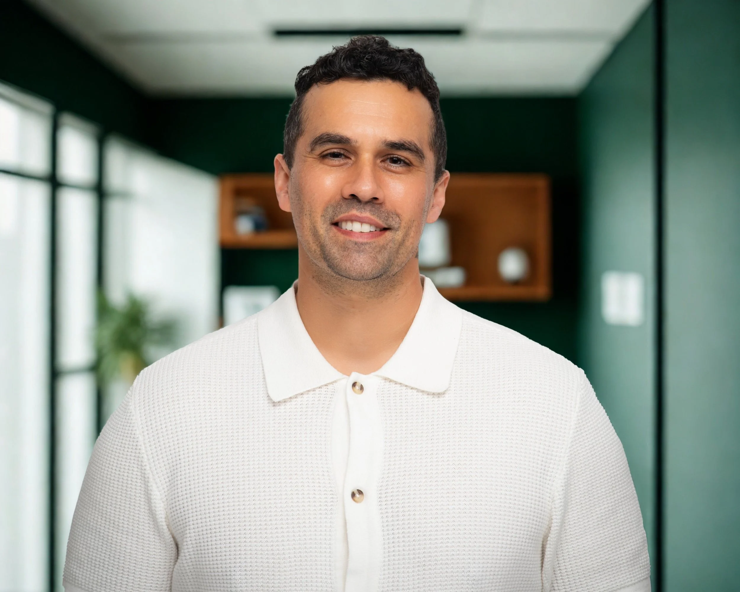 A smiling man with dark, curly hair, wearing a white textured polo shirt, standing in a room with green walls and large windows.