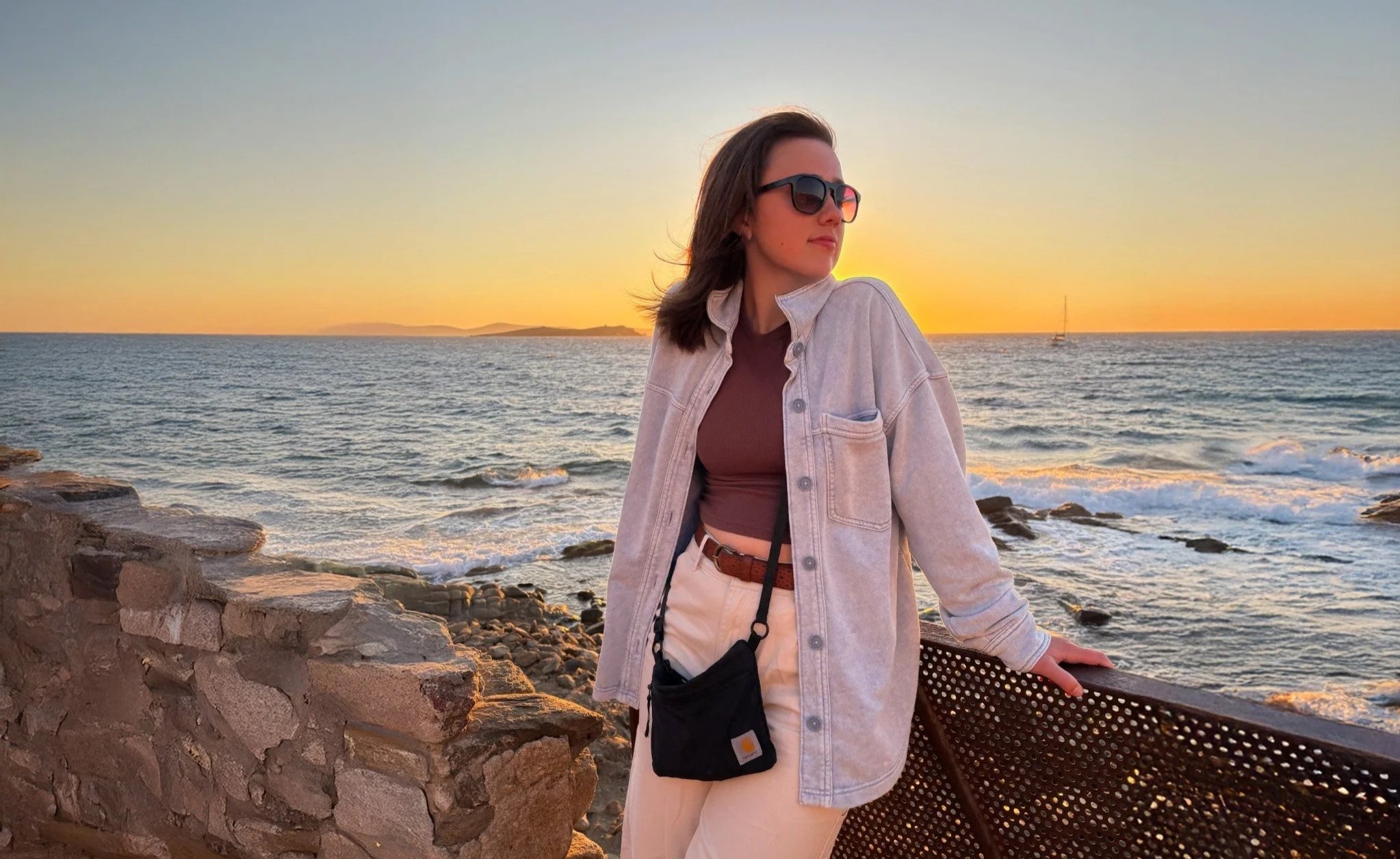 Sydney in sunglasses, beige jacket, brown top, and beige pants standing by ocean at sunset, with rocky shoreline in background.