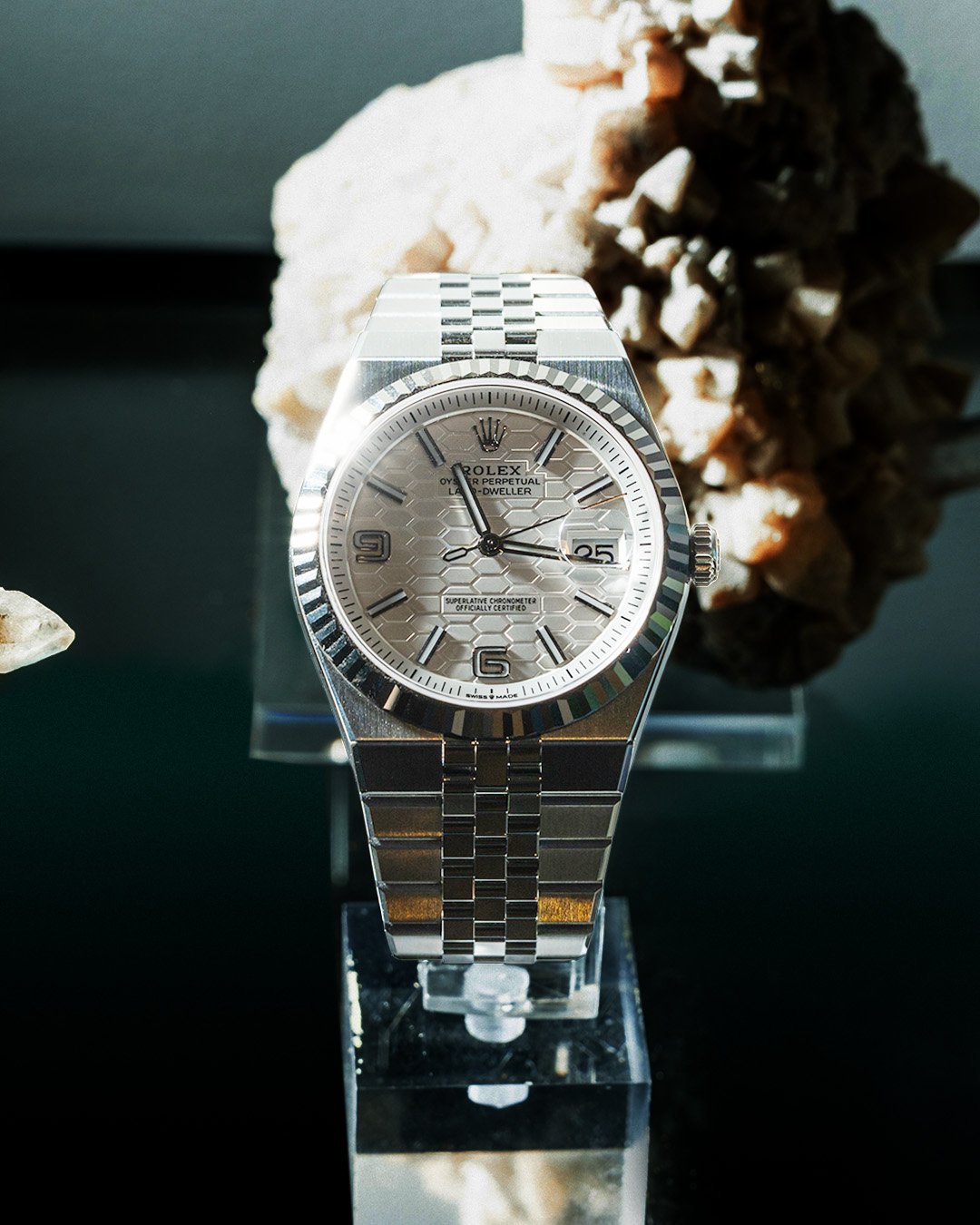 A silver Rolex watch with a honeycomb dial design, displayed on a transparent stand, with rocks in the background.