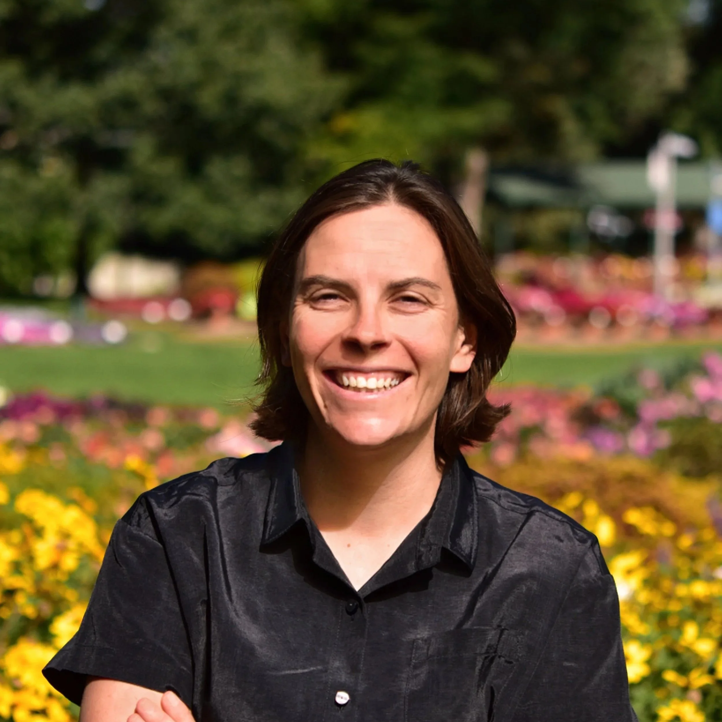 Close-up of a smiling woman with brunette hair, wearing a black top, standing outdoors with trees and colorful flowers in the background.