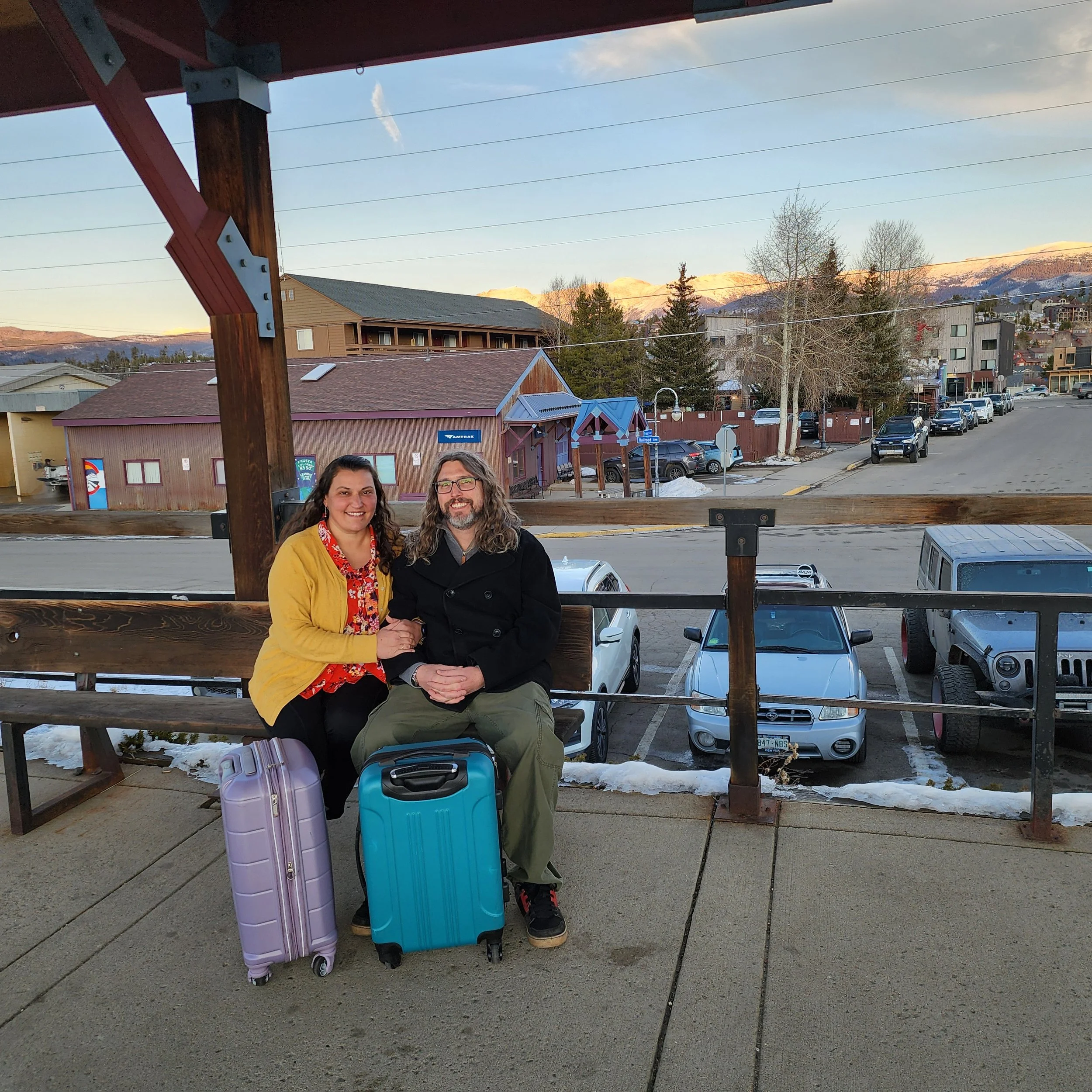 A smiling couple sitting on a wooden bench with purple and blue rolling suitcases, outdoors on a sidewalk, with a parking lot and small town buildings in the background during sunset. Suitcase, Ski bag, Snowboard tote, luggage,