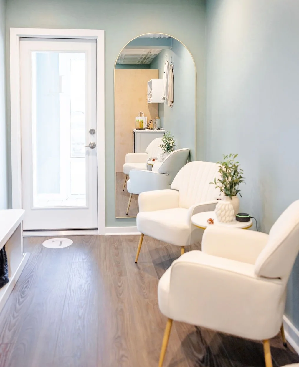 A cozy waiting area with three white armchairs with wooden legs, a round white side table with a potted plant, and a large mirror on a light blue wall. A door with glass panels is visible on the left.