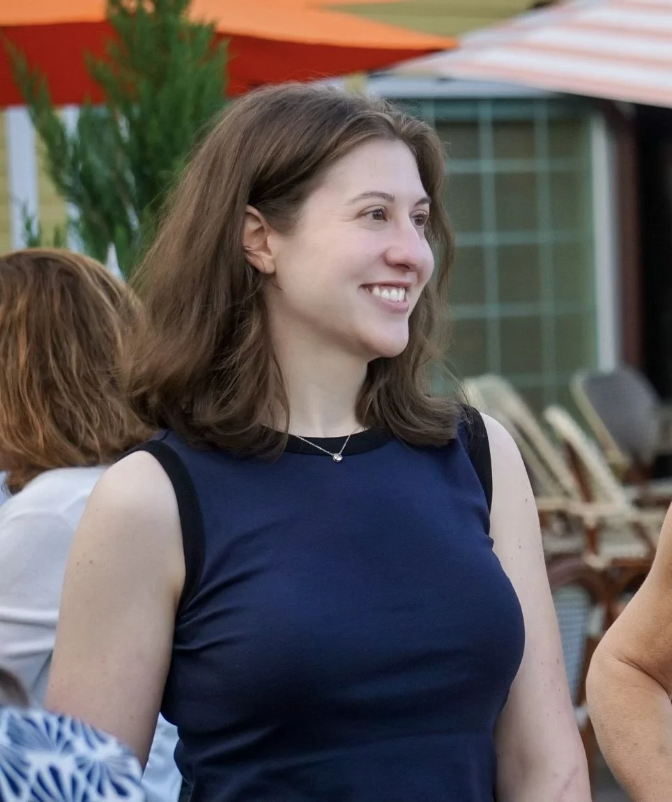 A young woman with shoulder-length brown hair smiling outdoors during daytime, wearing a navy sleeveless shirt and a necklace, with colorful umbrellas and chairs in the background.