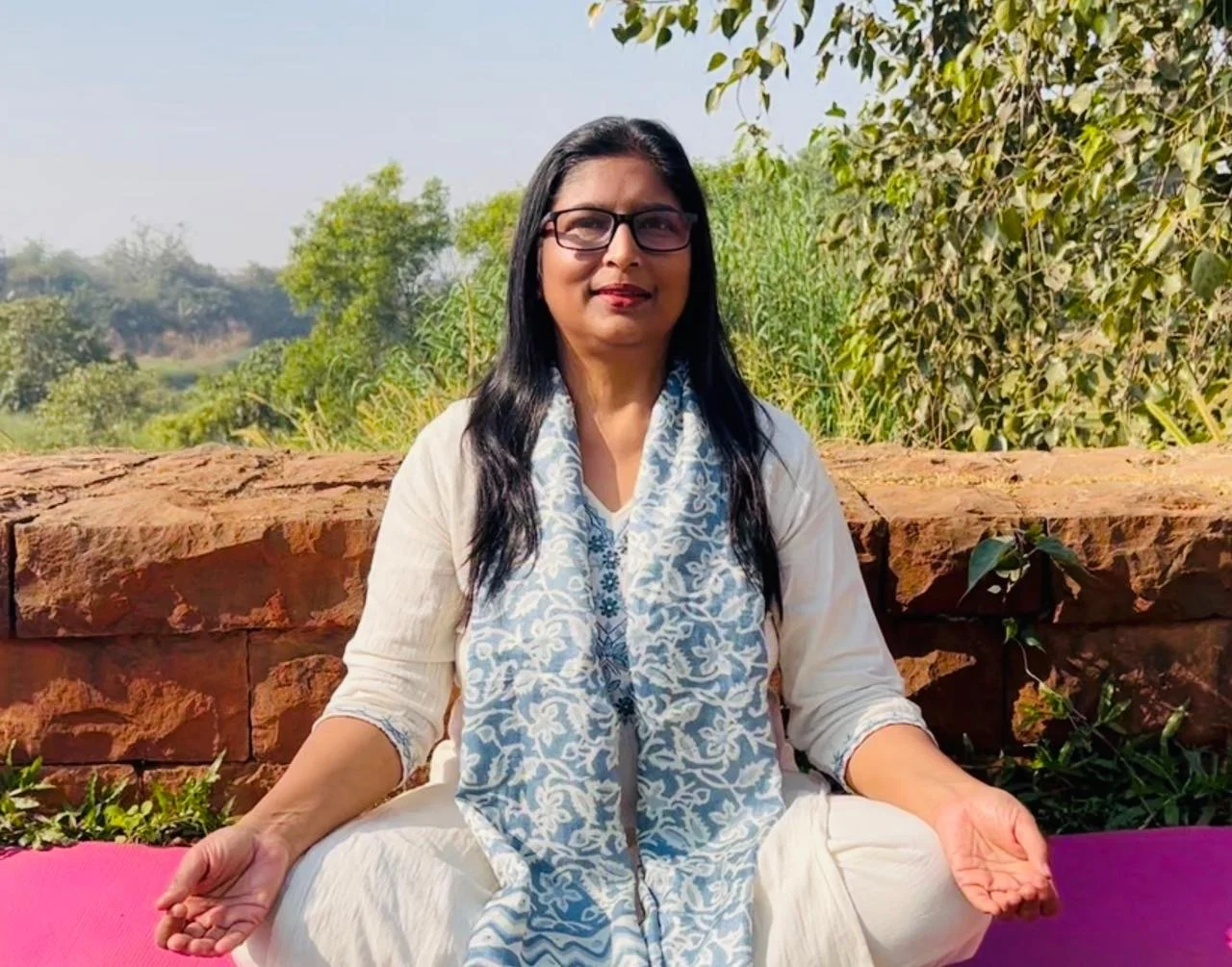 Woman practicing yoga outdoors, sitting cross-legged on a pink mat, with greenery and trees in the background.