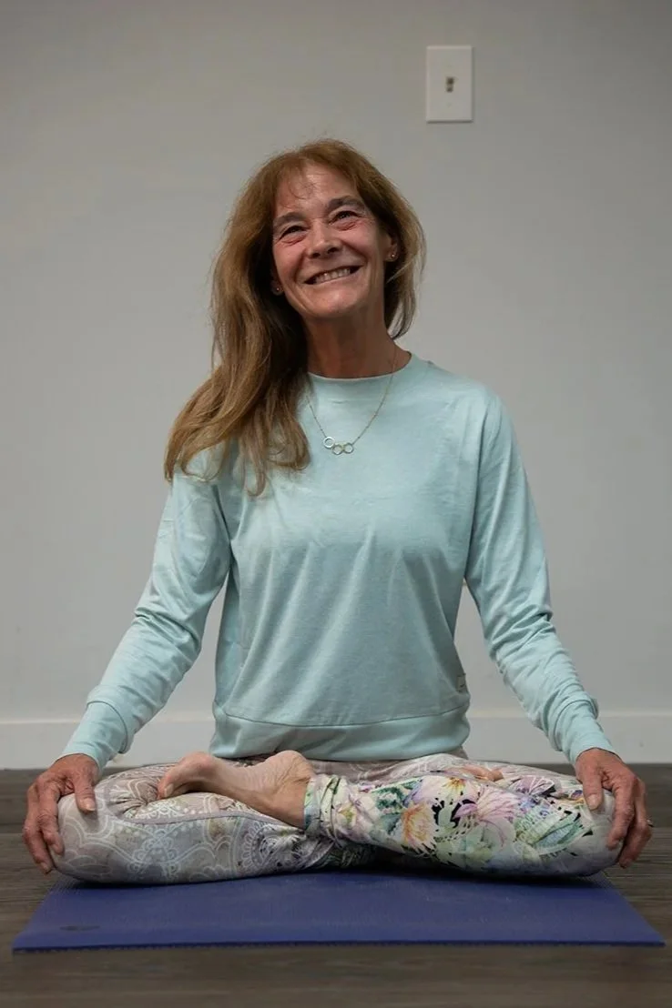 A woman with long hair and a big smile practicing yoga on a mat, sitting cross-legged with one foot resting on her thigh in a peaceful indoor setting.