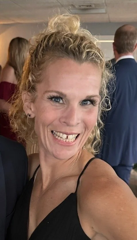 A woman with curly blonde hair smiling at the camera, wearing a black spaghetti strap top, at a social event with people in formal attire in the background.