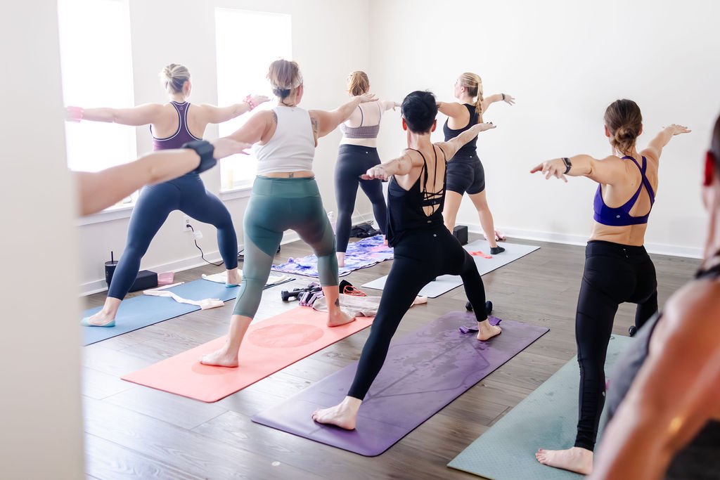 Group of women participating in a yoga class practicing Warrior pose in a bright studio.