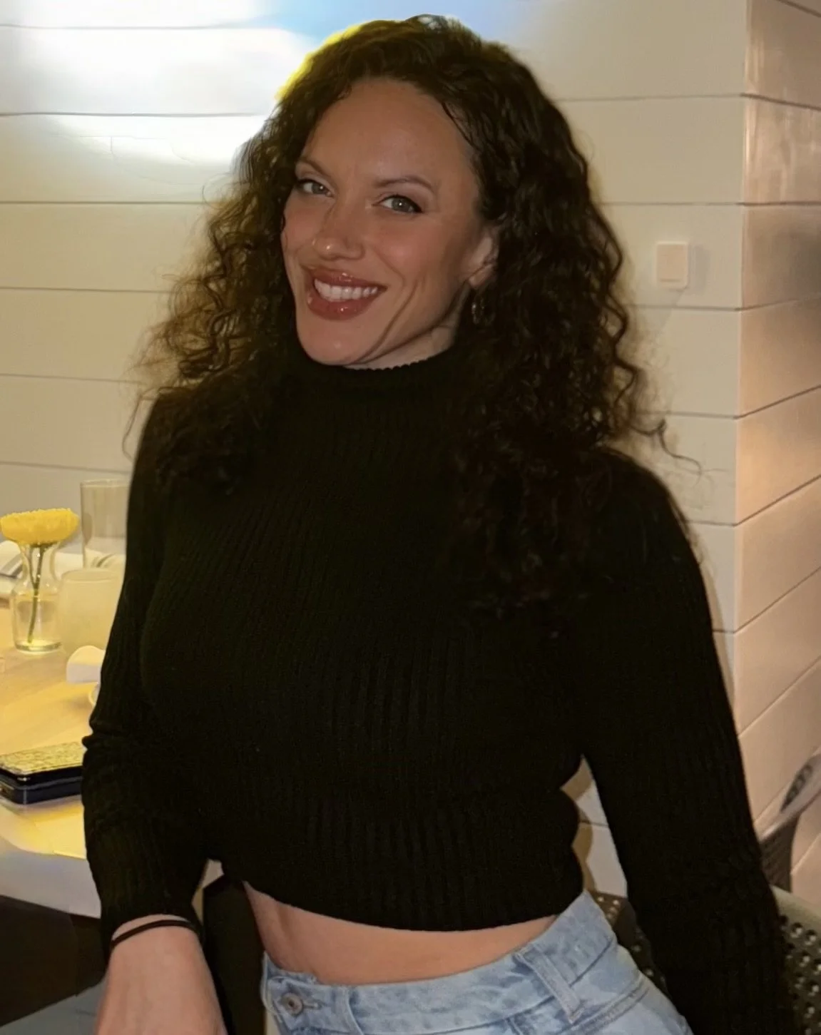 A woman with curly dark hair smiling in a black long-sleeve top and light blue jeans, standing indoors near a white paneled wall.