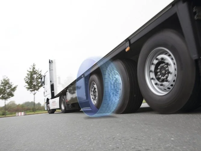Moving semi-truck trailer with blue digital tire sensor or camera on the front tire, on a road with trees and overcast sky. Truck tyre technology.