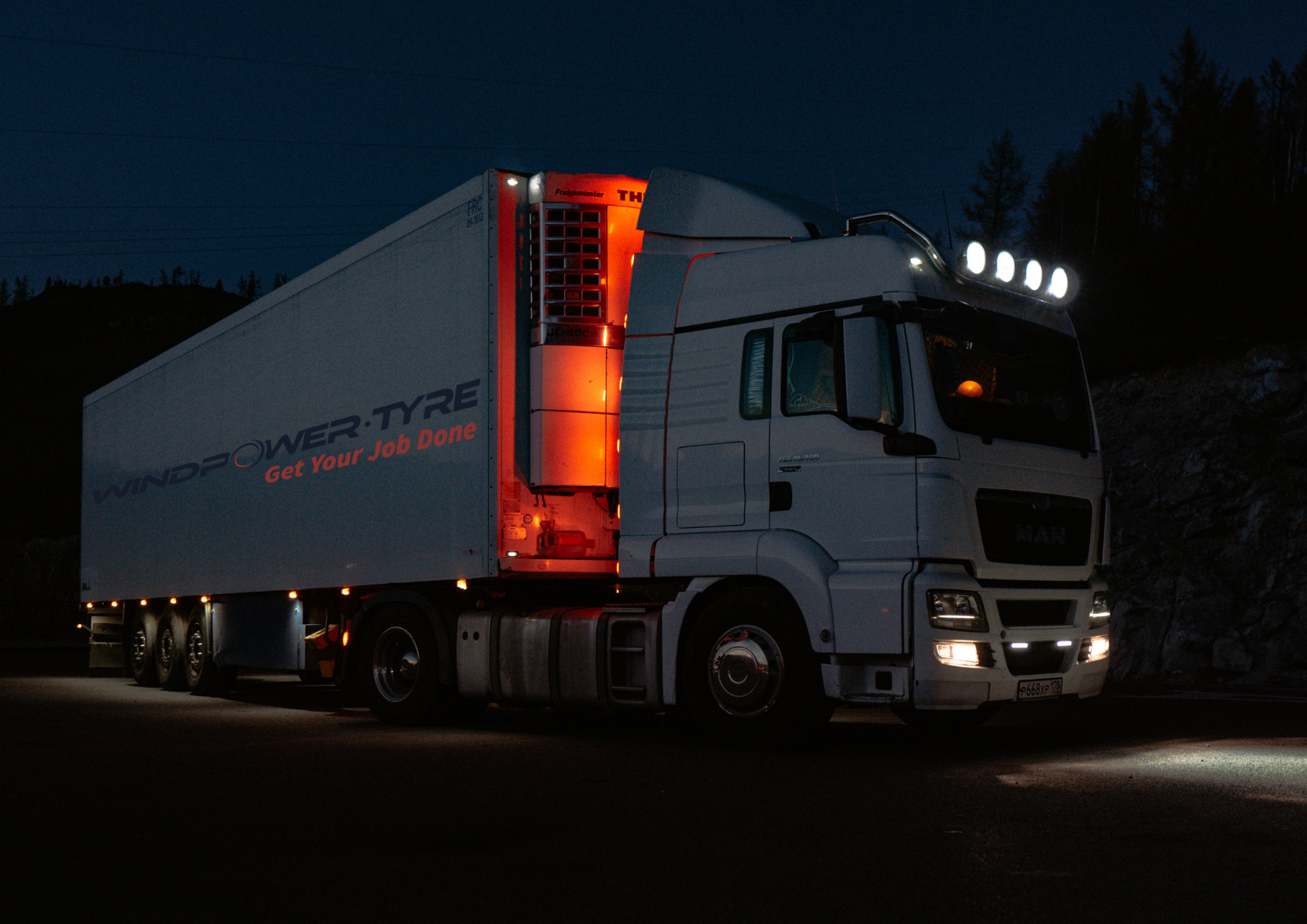 A large white truck with the brand name 'MAN' visible on the front, parked on a dark road at night, with lights on the roof and side, and a logo on the side that reads 'WINDPOWER TYRE' and 'Get Your Job Done'.