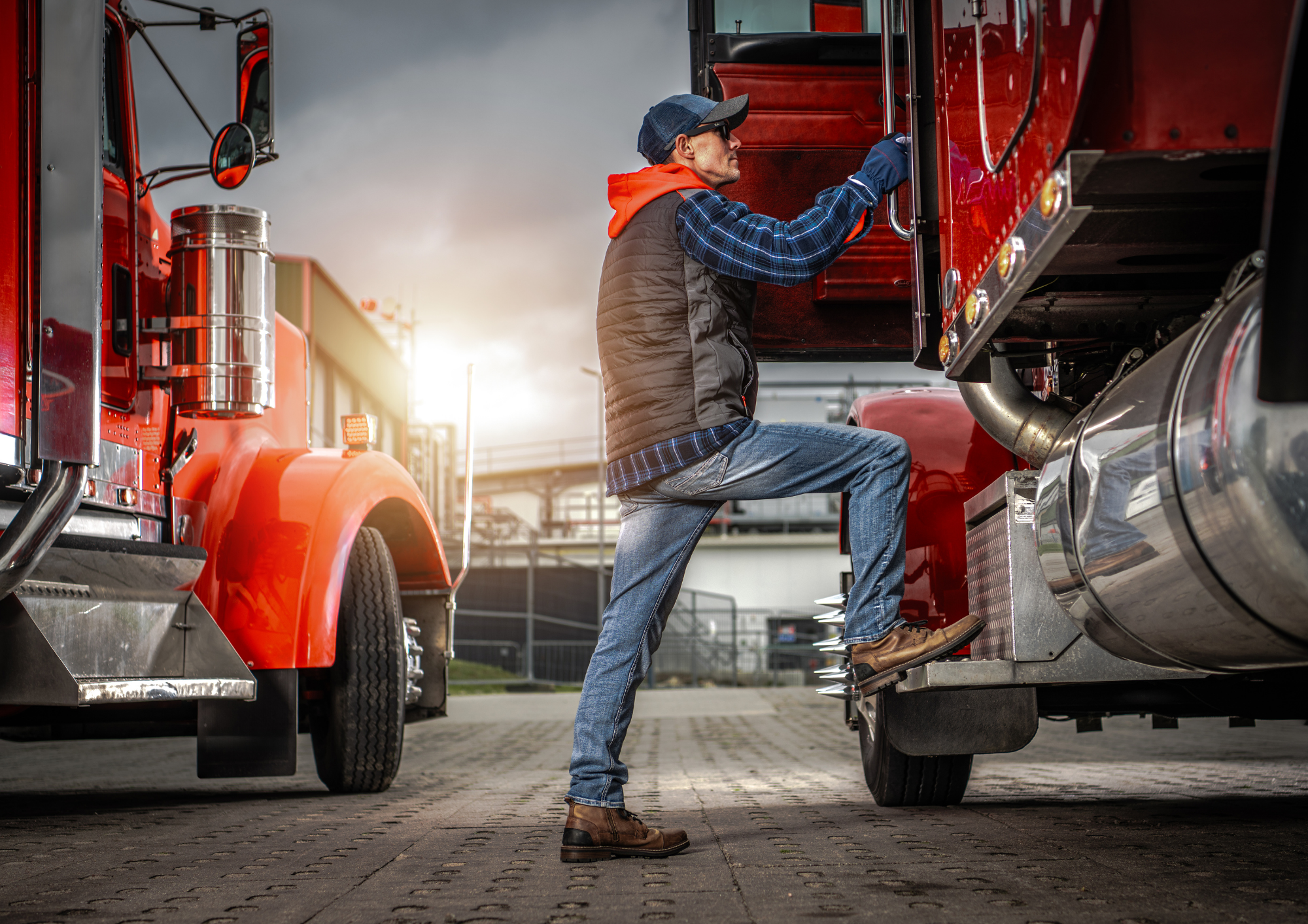 A man in a cap, sunglasses, and a plaid shirt, kneeling with one foot on a step of a large red fire truck, reaching into an open compartment as the sun setting in the background. Truck Tyres & TBR Tyres.