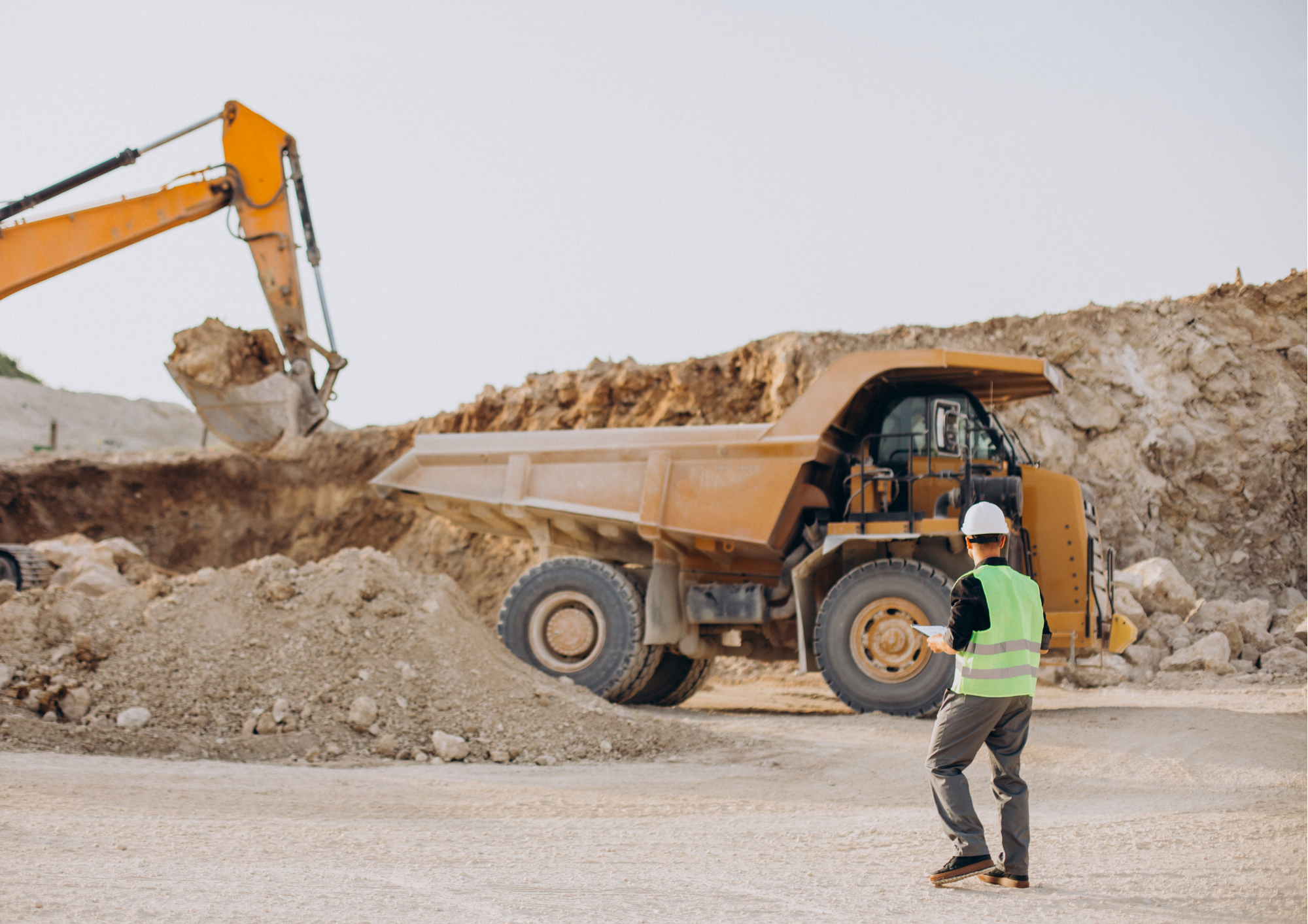 Construction site with a yellow dump truck and excavator, worker in safety vest and helmet holding a tablet. Earthmover Tyres. Earthmoving Tyres.