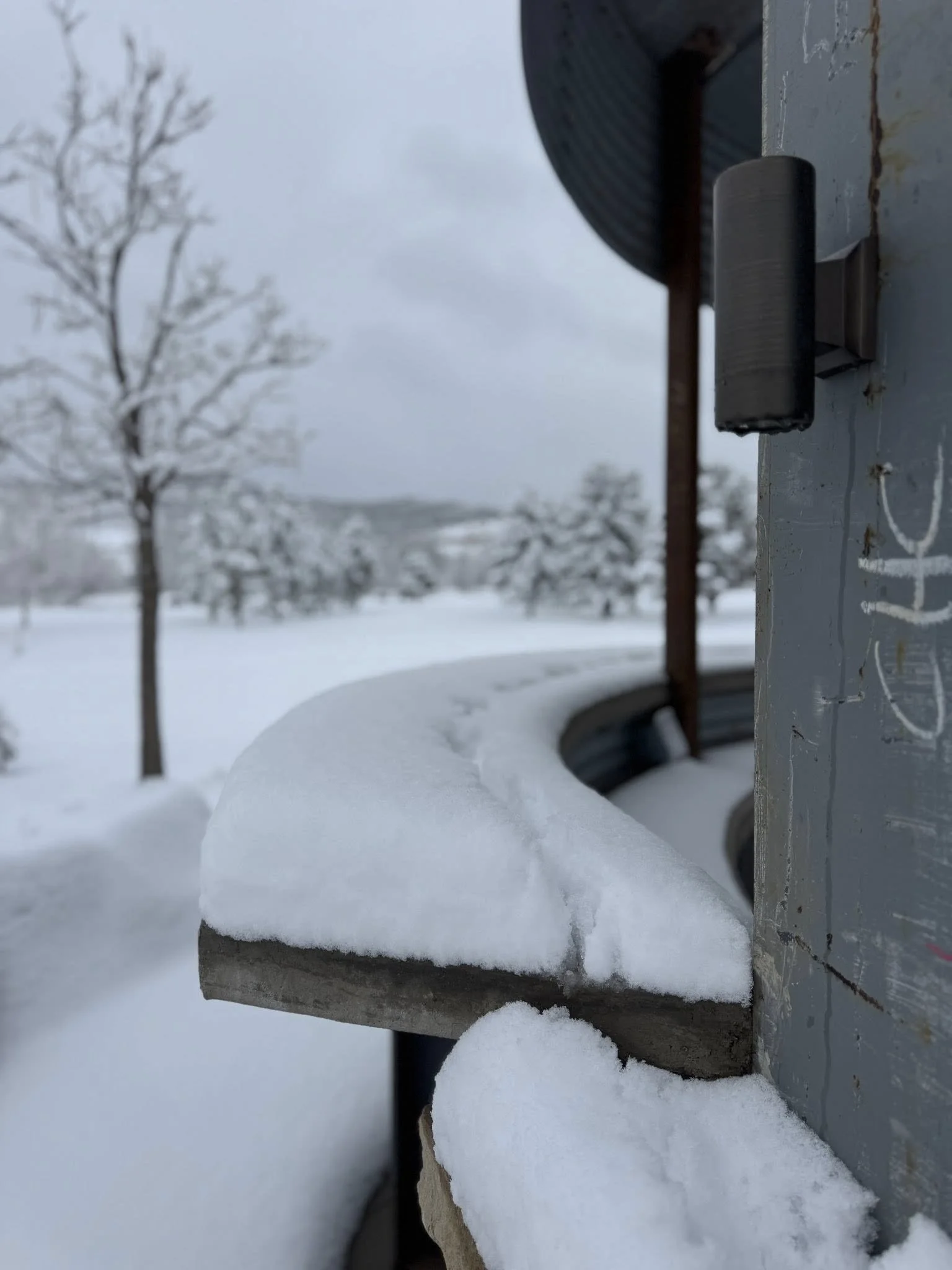 Close-up of a snow-covered ledge on a utility box with a snowy landscape and leafless trees in the background.
