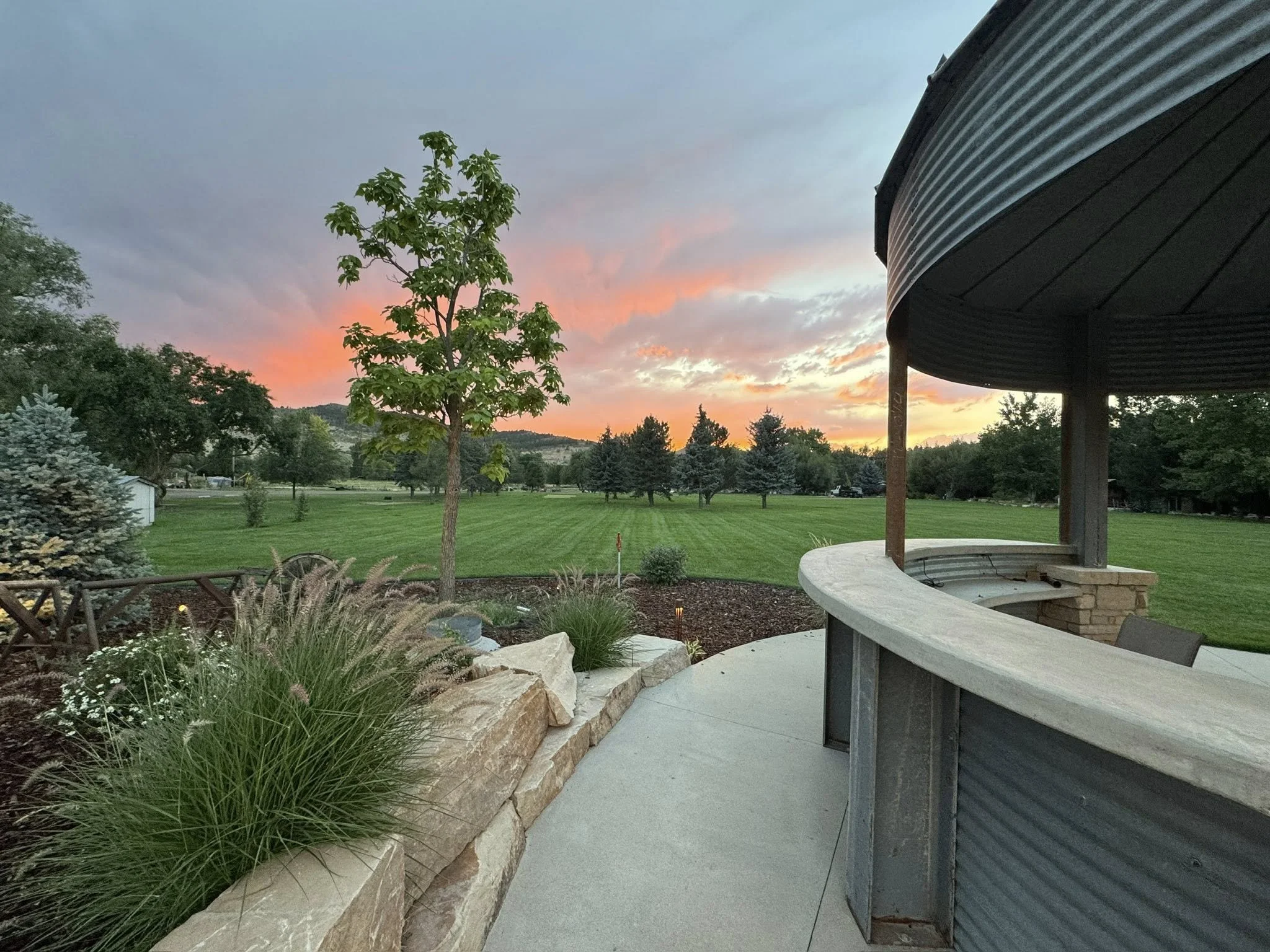 A scenic outdoor view at sunset with a curved concrete patio, a metal gazebo, trees, and a large grassy field.