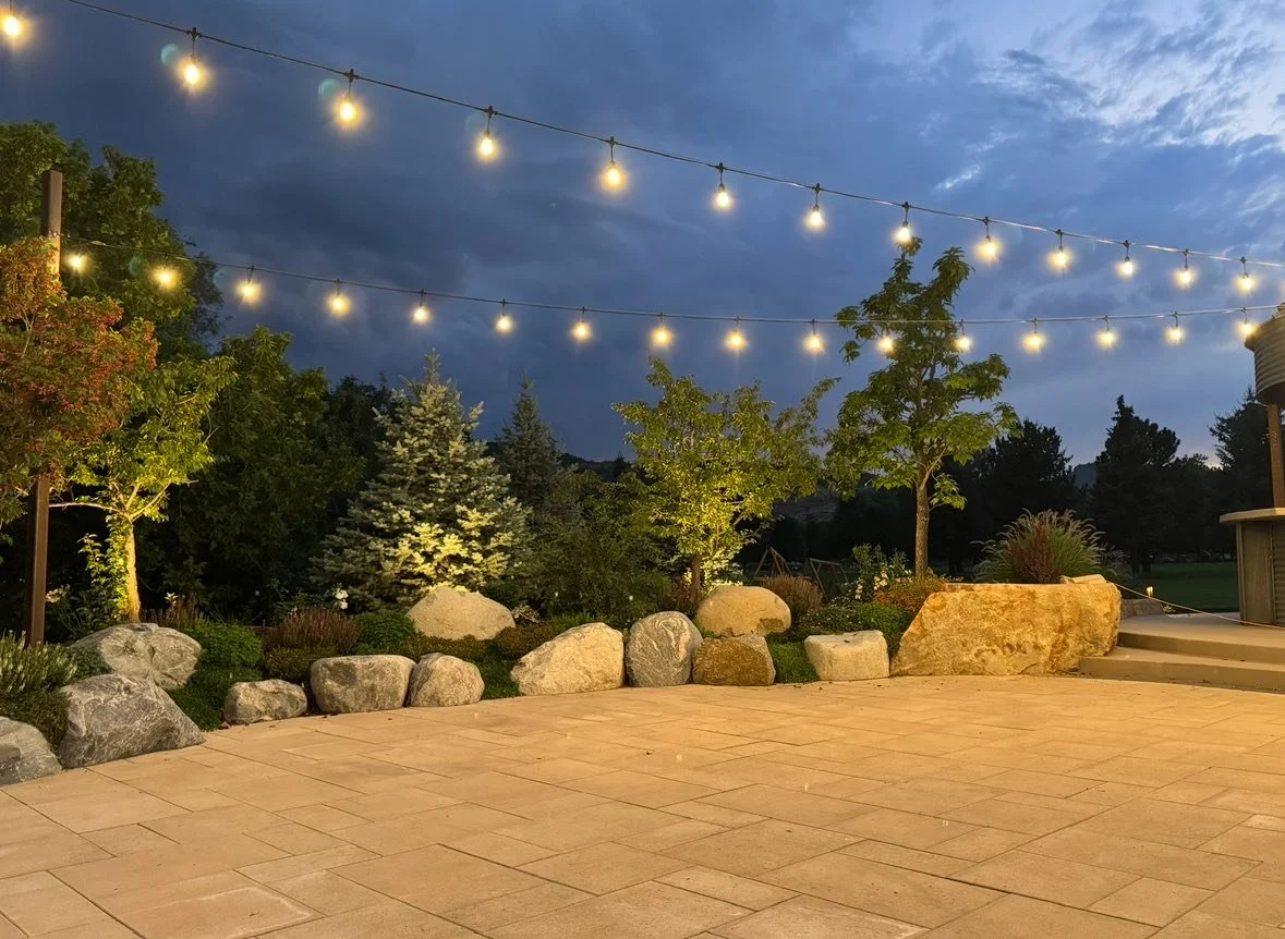 A backyard patio scene at dusk with string lights hanging overhead, trees, rocks, and a clear evening sky.