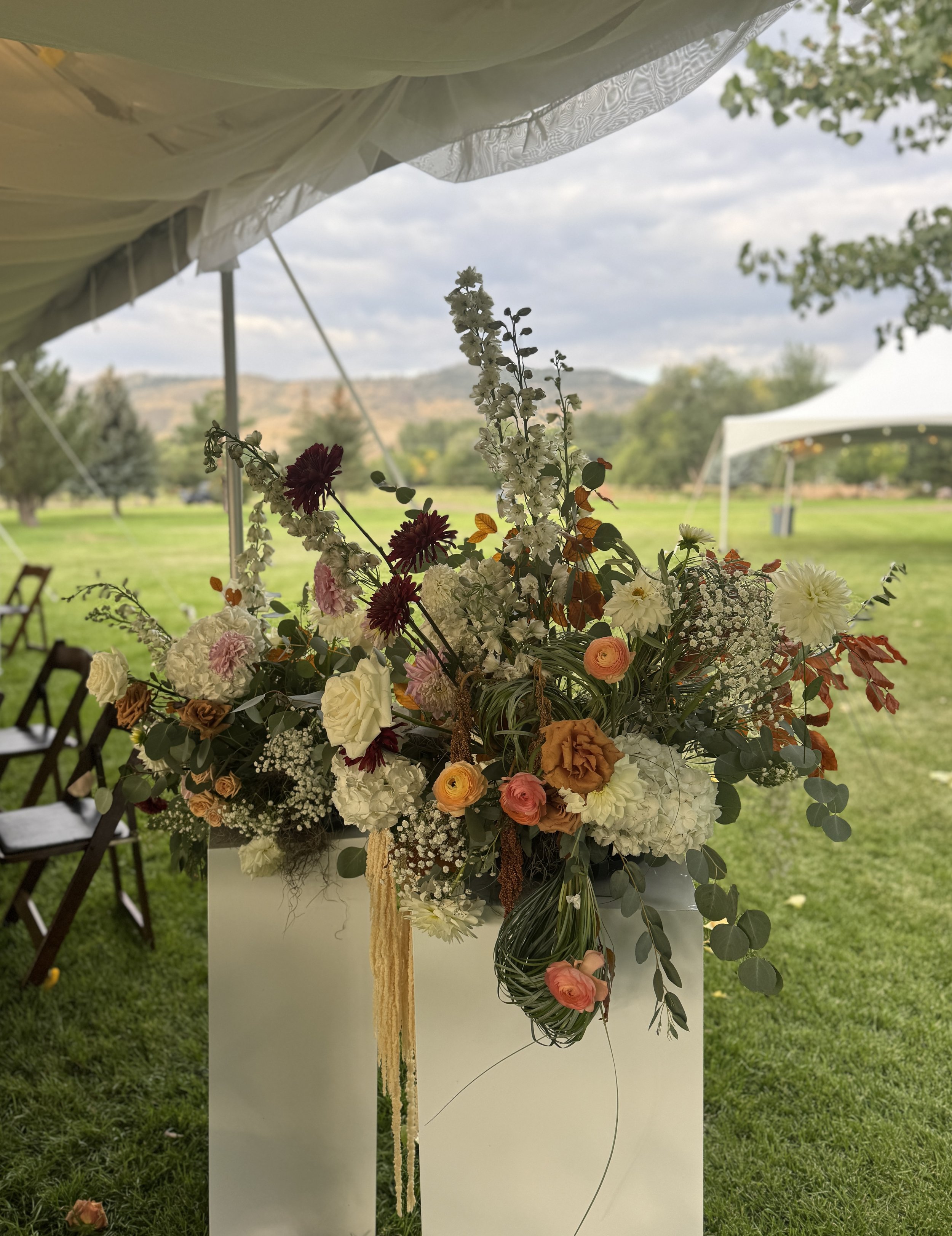 A floral arrangement with white, pink, orange, and burgundy flowers, including hydrangeas, roses, dahlias, and other blooms, displayed on a white pedestal outdoors under a tent, with chairs and a grassy field in the background.