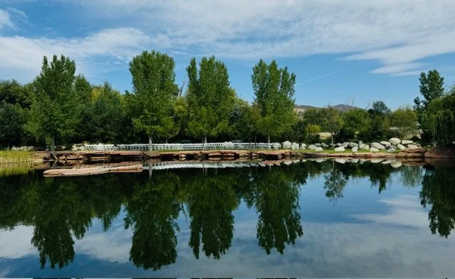 Calm river reflecting green trees and a blue sky with clouds, rocky shoreline with a white fence in the background.
