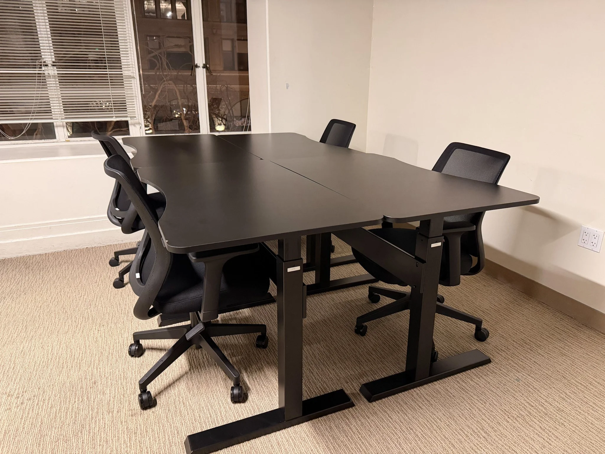 An empty conference room with a large black table and six black office chairs around it, with a window in the background and beige carpeted floor.