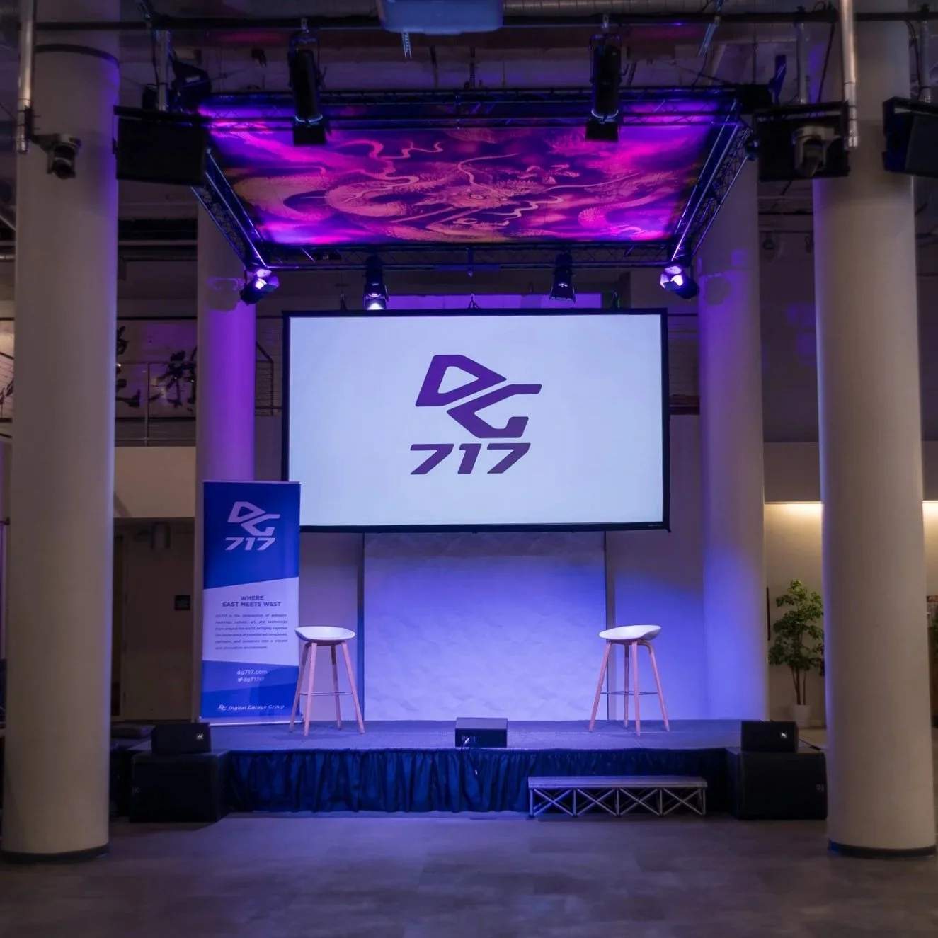 Stage with two white stools, a large screen displaying a logo, and a vertical banner with the logo and text, in a dimly lit venue with purple lighting and large white columns.