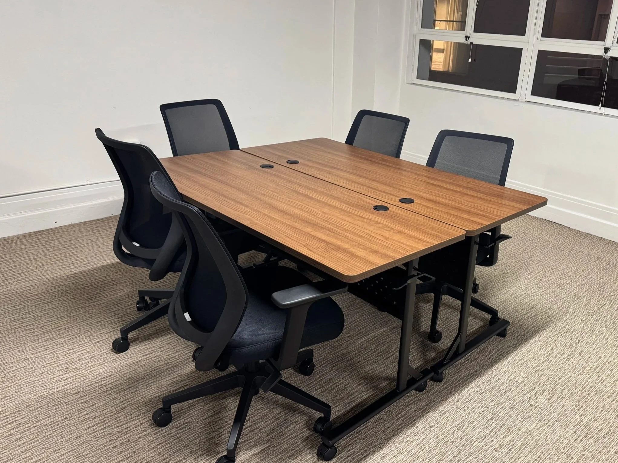 Empty conference room with a wooden table and six black and gray office chairs, with windows and beige carpet.