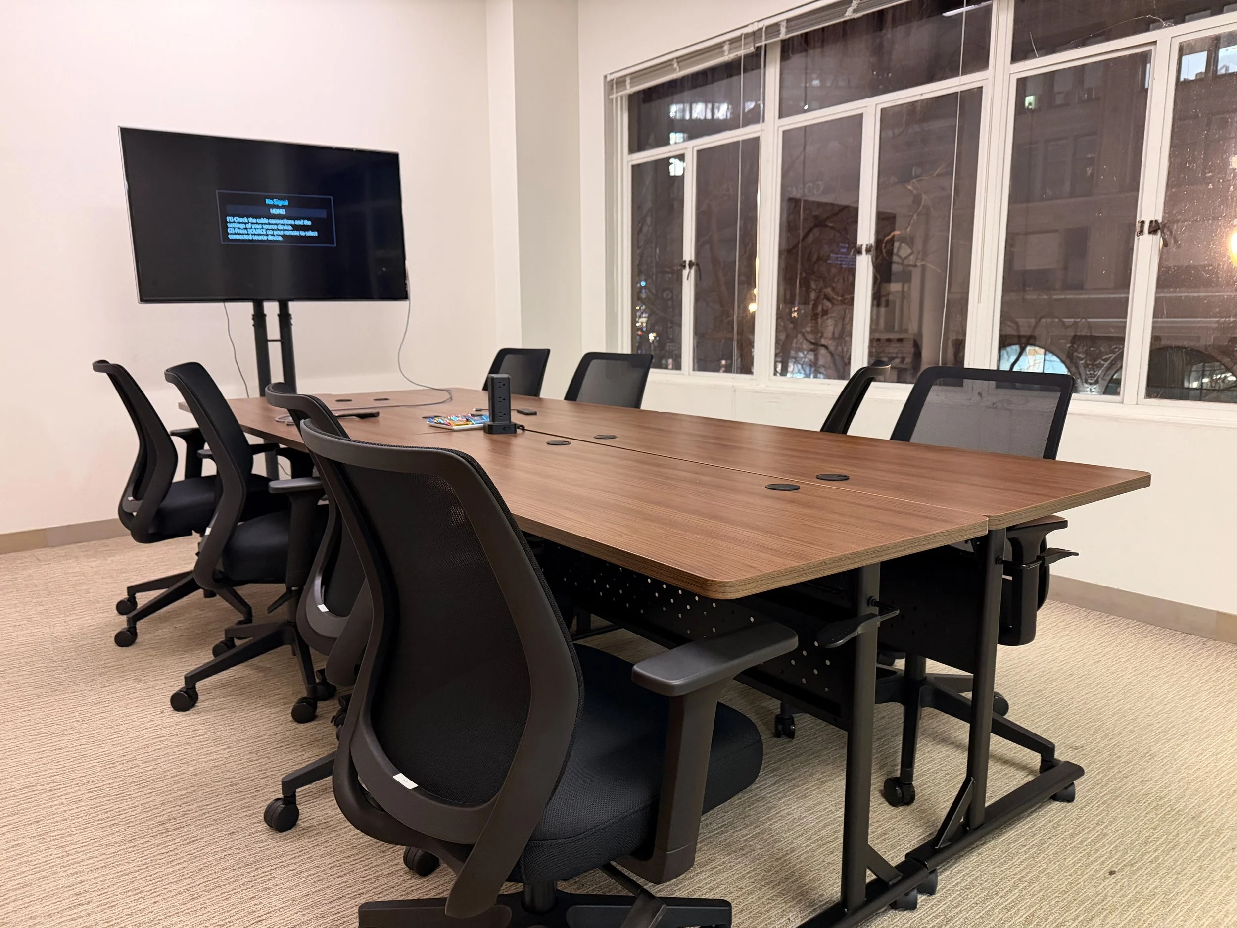 Empty conference room with a wooden table and black ergonomic chairs, a large window behind the table, and a TV screen on the wall showing no signal.