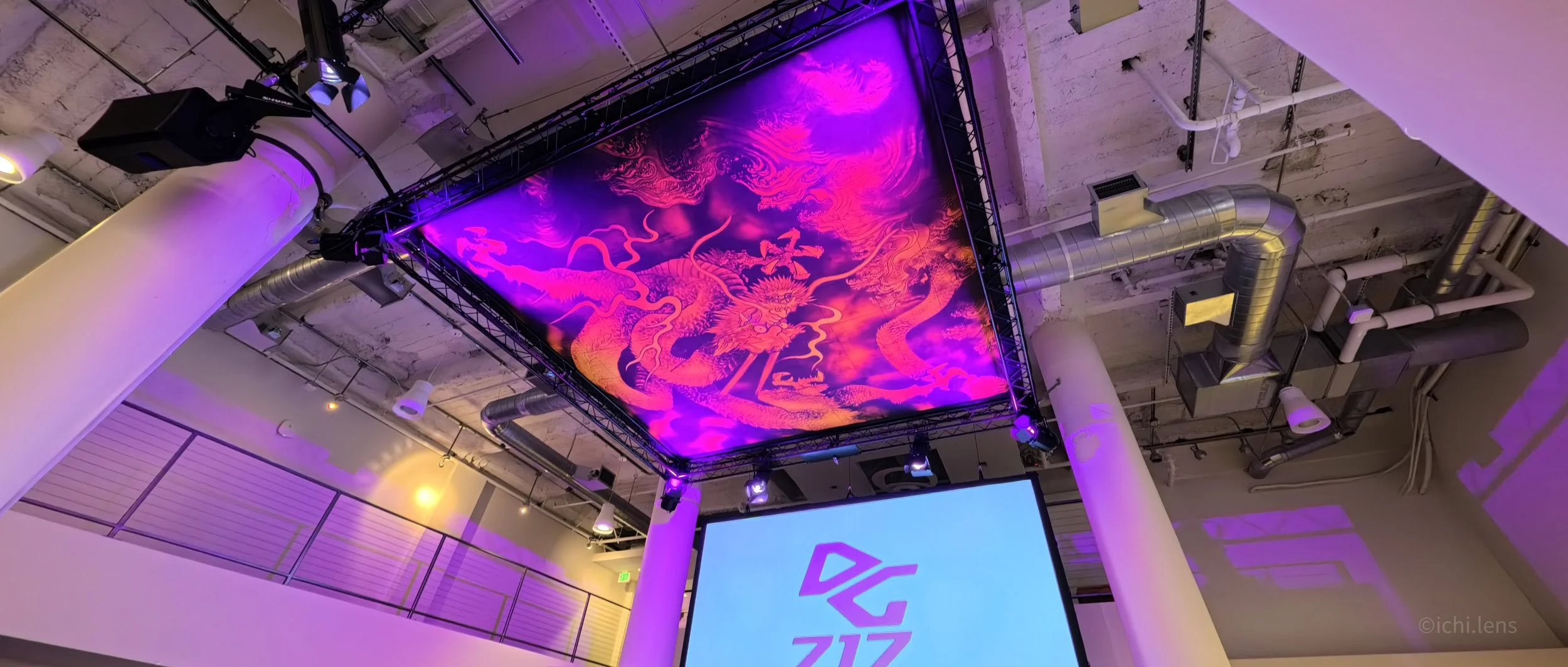 Interior of a modern space with a large illuminated LED screen displaying a dragon on a purple and orange background, hanging from a metal frame on the ceiling. Exposed ductwork and industrial lighting are visible on the ceiling.