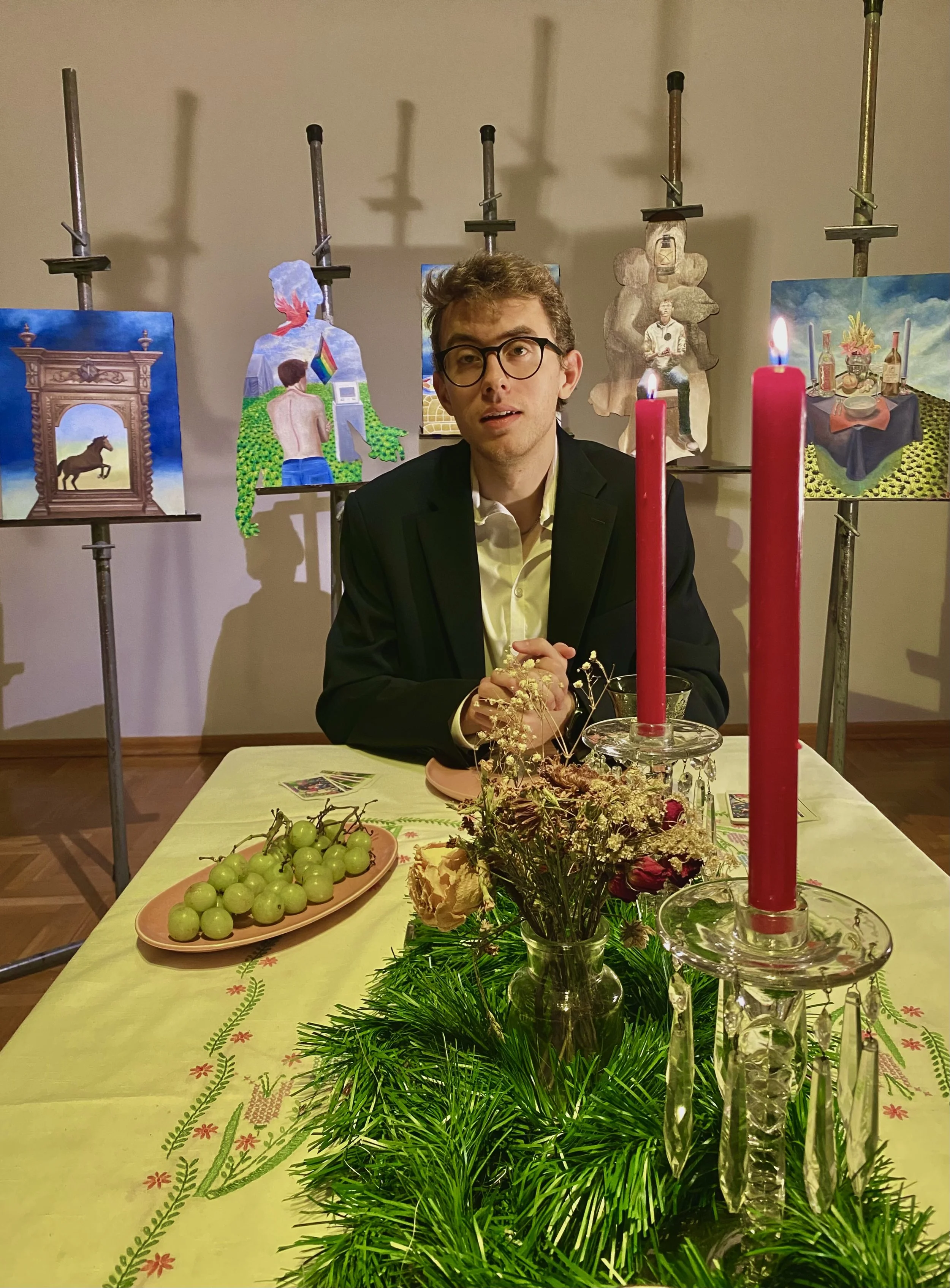 A man in glasses and a black blazer is sitting at a decorated table with red candles in glass candle holders, a plate of green grapes, and a floral centerpiece. Behind him, paintings are displayed on easels, and the room has a simple beige wall.