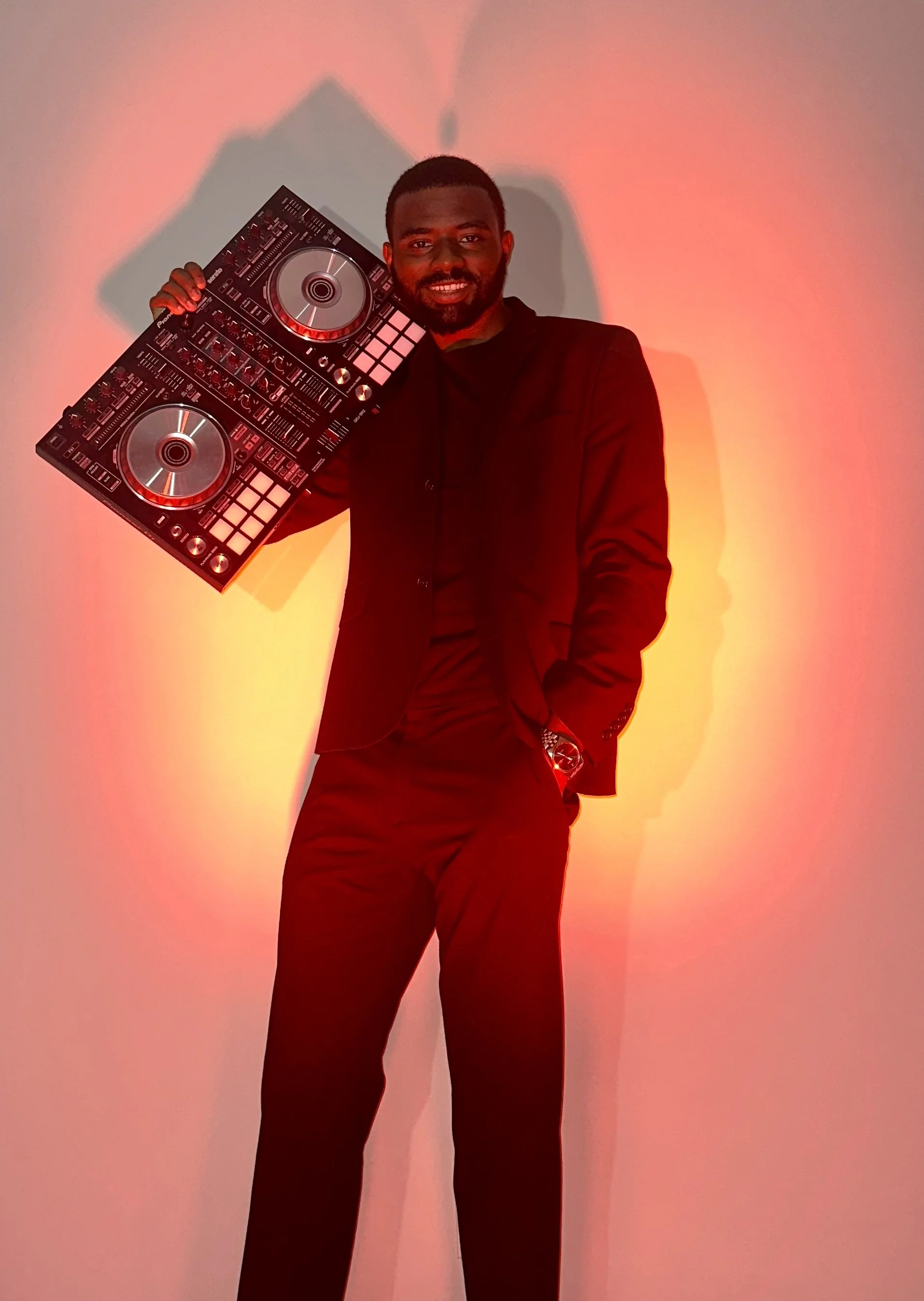 A man in a black suit holding a DJ controller, standing against a pink and yellow colored wall.