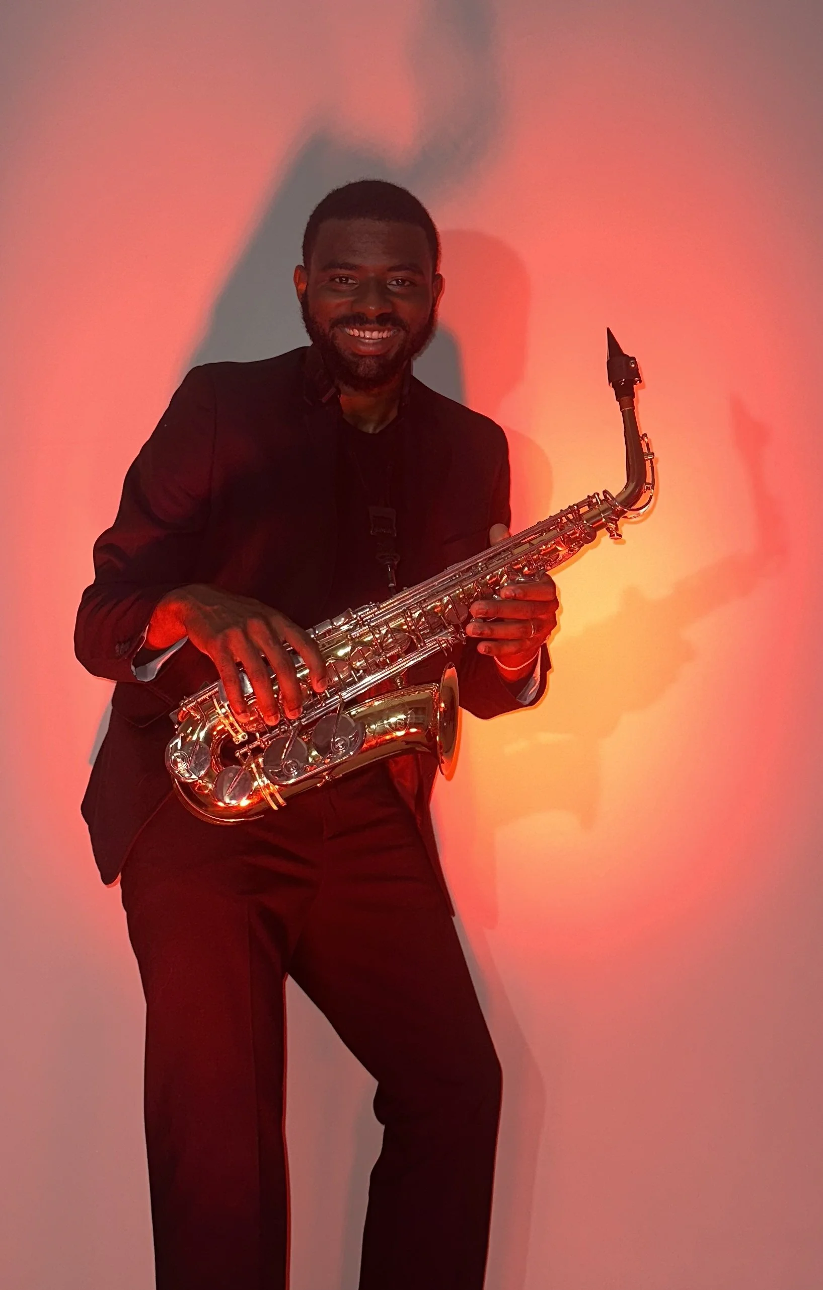 Man in black suit playing a shiny saxophone against a red background.