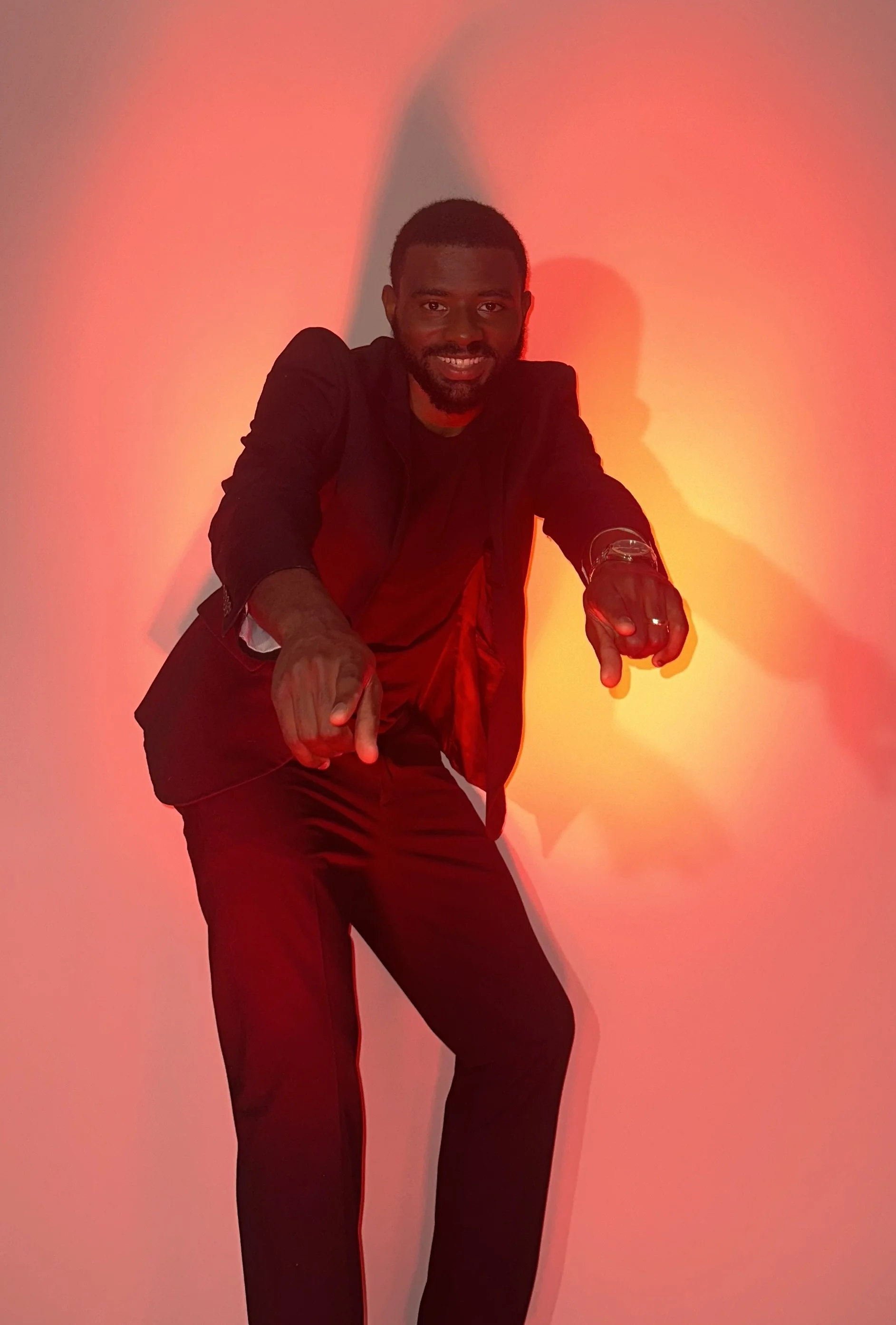 A man with dark skin and a beard, dressed in black, pointing towards the camera with a smile, standing against a vibrant red and orange lighting backdrop