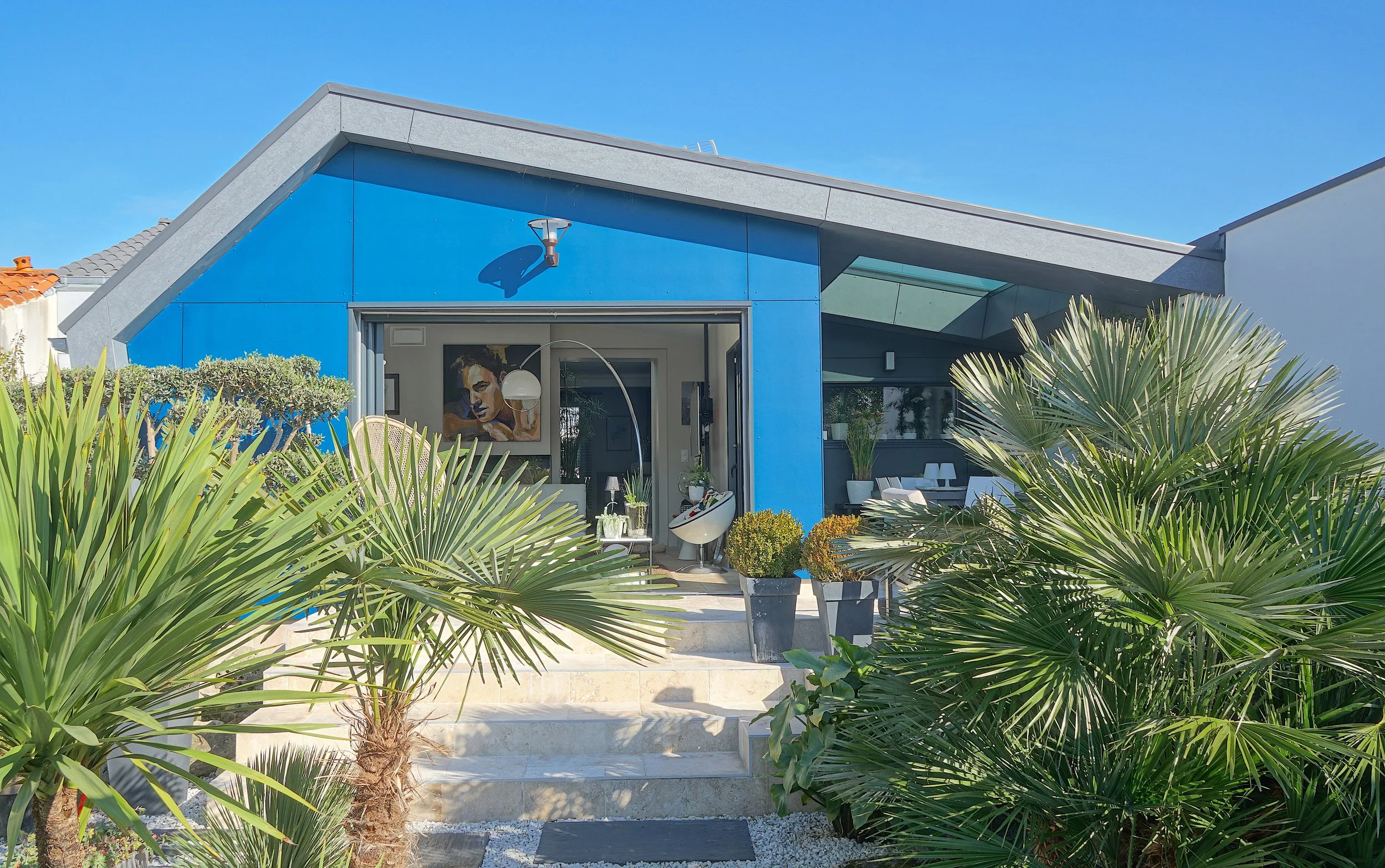 Une maison moderne avec une façade bleue, un jardin avec des palmiers et des pots de fleurs, et un intérieur visible avec un tableau de portrait et des meubles blancs.