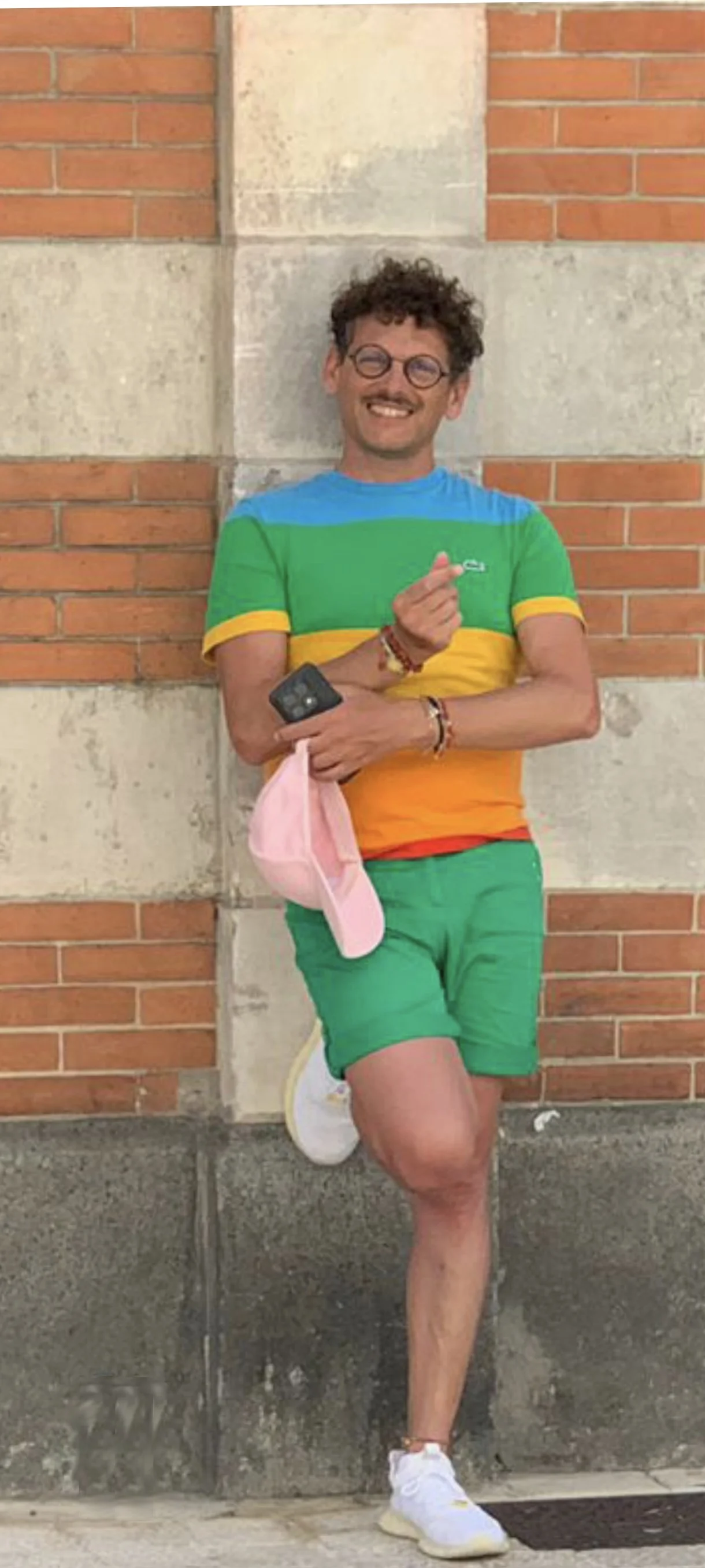 Jeune homme souriant, vêtu d'un t-shirt rayé vert, bleu, jaune et orange, portant des lunettes rondes, un bracelet, des chaussures blanches, debout contre un mur en briques et pierre.