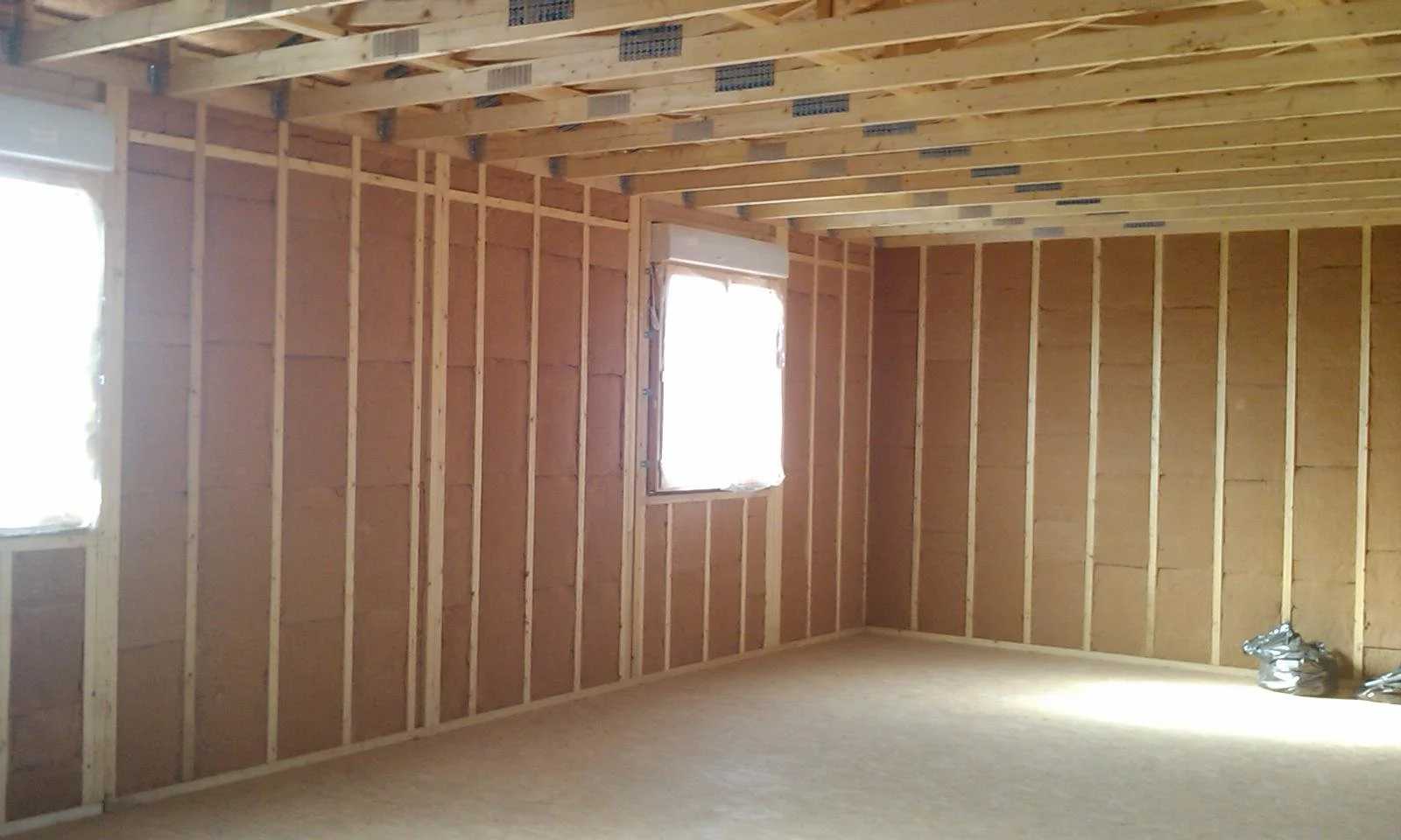 Une pièce en construction avec murs et plafond en bois, sans finition, avec deux fenêtres. Sol en bois ou en revêtement semblable, et des outils ou matériel au coin droit.