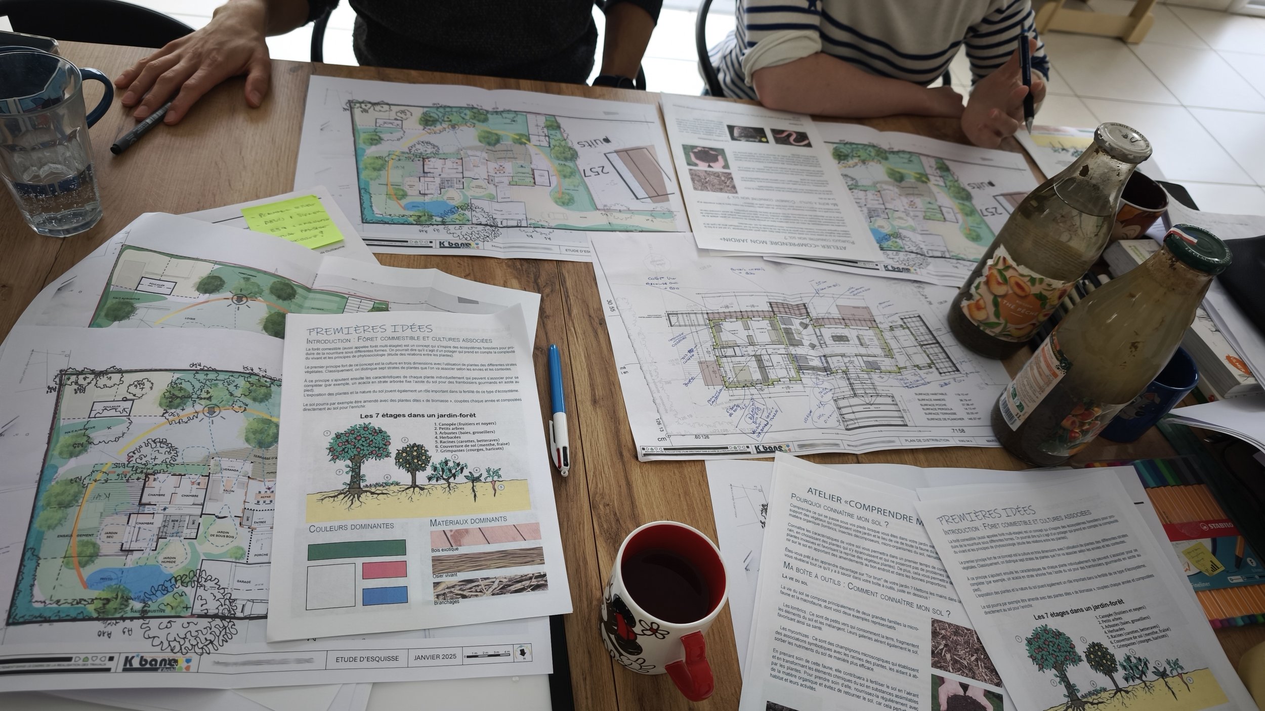 Table de travail avec plans d'architecture, documents, dessins, stylos, deux bouteilles, tasse de café, et personne qui écrit.