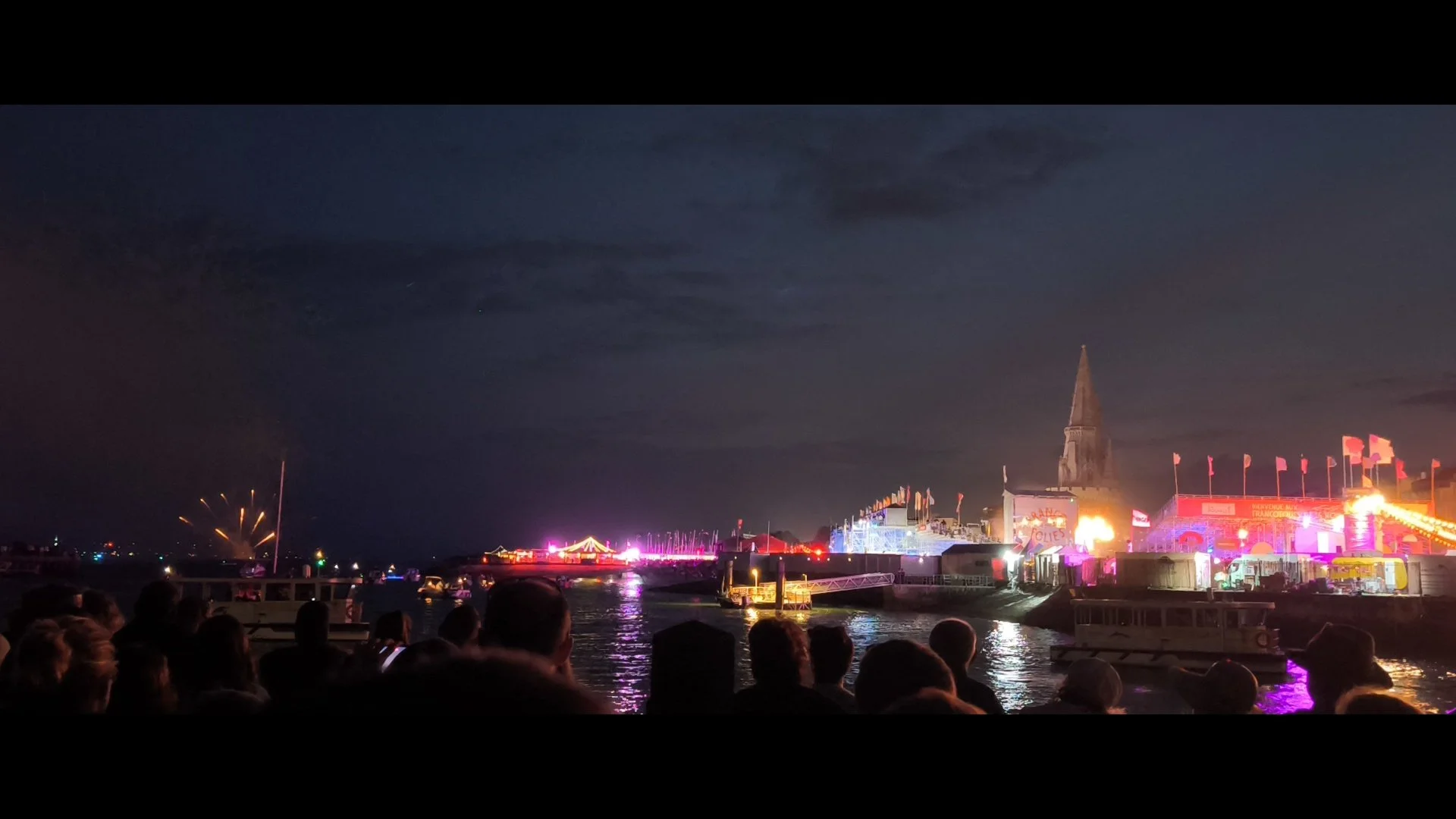 Célébration nocturne au bord de l'eau avec feux d'artifice, foule assise, structures illuminées, et un grand bâtiment avec un clocher pointu en arrière-plan.