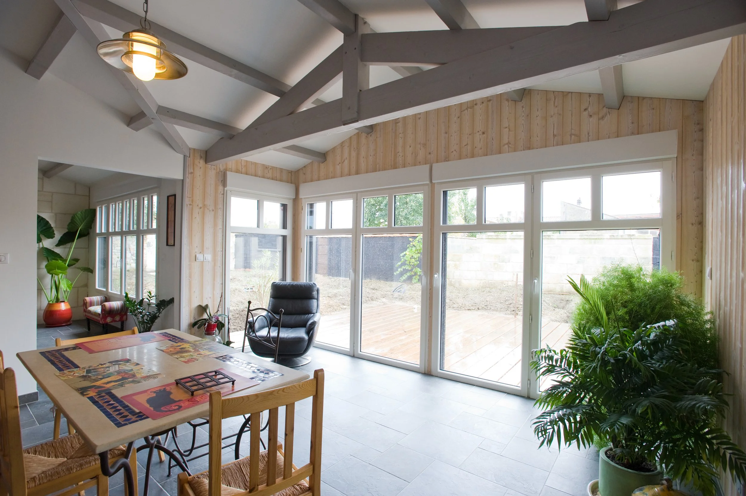 Intérieur lumineux d'une salle à manger avec grande baie vitrée, plantes vertes, table en bois avec décoration, fauteuil noir, plafond avec poutres en bois peintes en blanc.