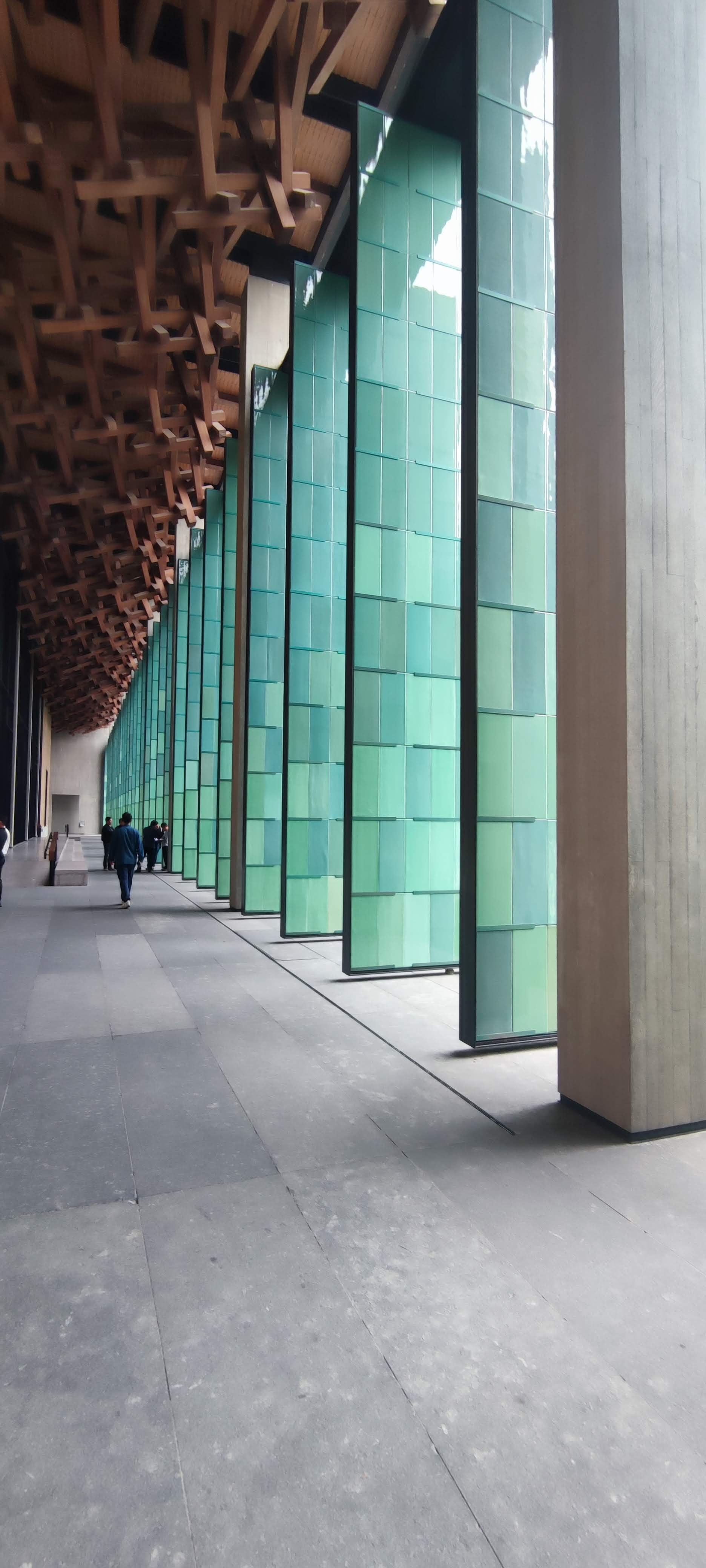Vue intérieure d'un bâtiment moderne avec de grandes fenêtres en verre vert, des colonnes en béton et un plafond en bois avec une structure complexe en bois massif.