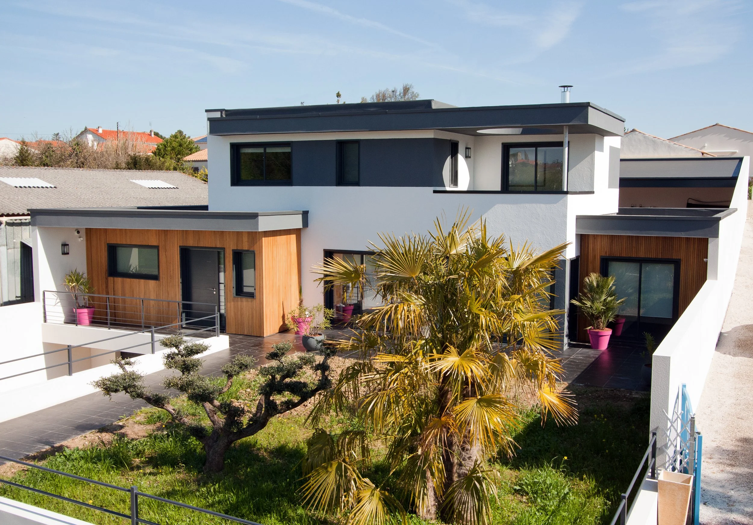 Maison bois contemporaine à deux étages avec façade en bois et blanc, jardins avec palmiers et arbres taillés, autour d'une clôture en métal, sous un ciel clair.