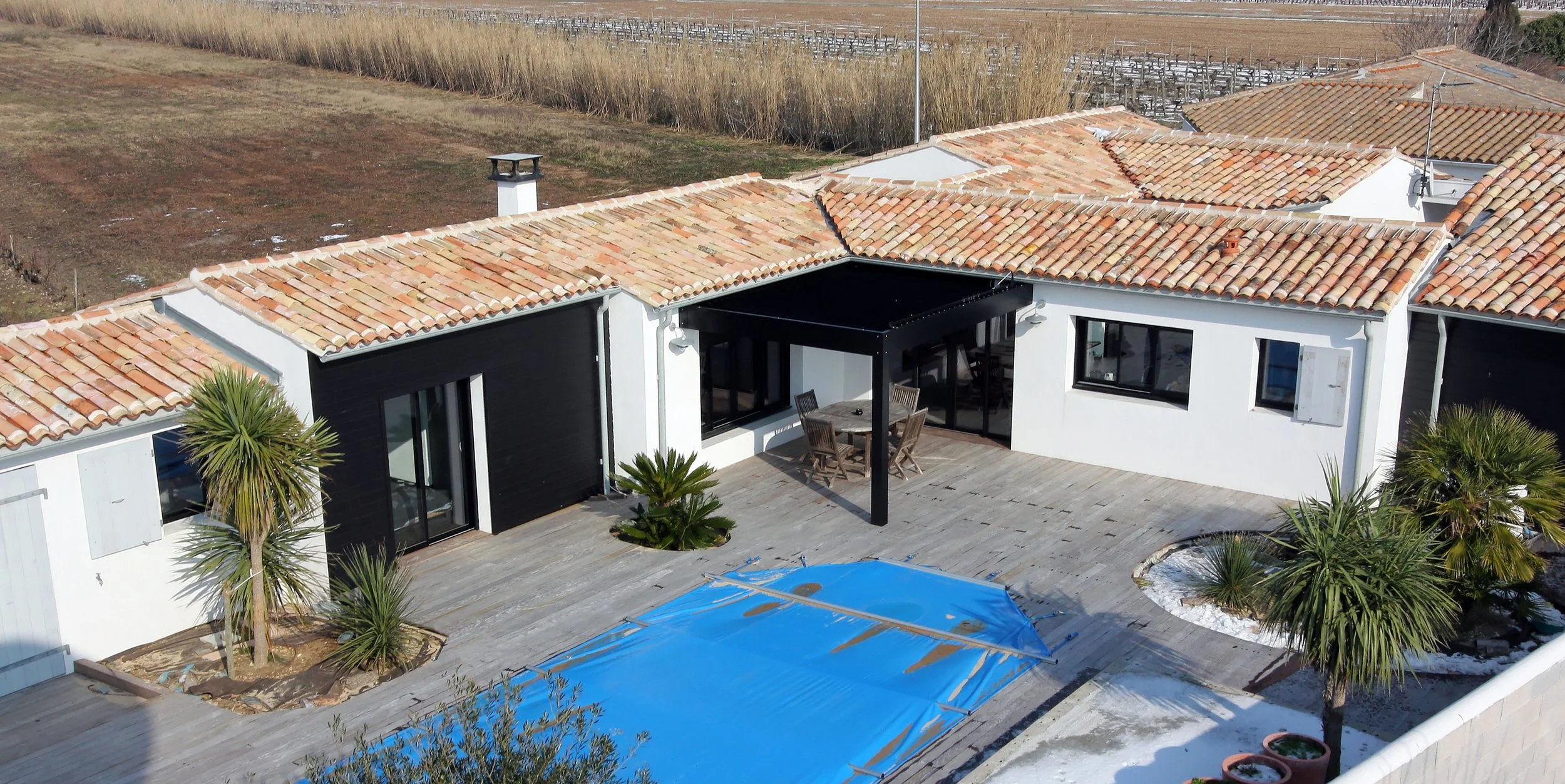 Vue aérienne d'une terrasse de maison avec piscine couverte par une bâche bleue, entourée de palmiers, avec toit en tuiles rouges, et un mobilier d'extérieur en bois.