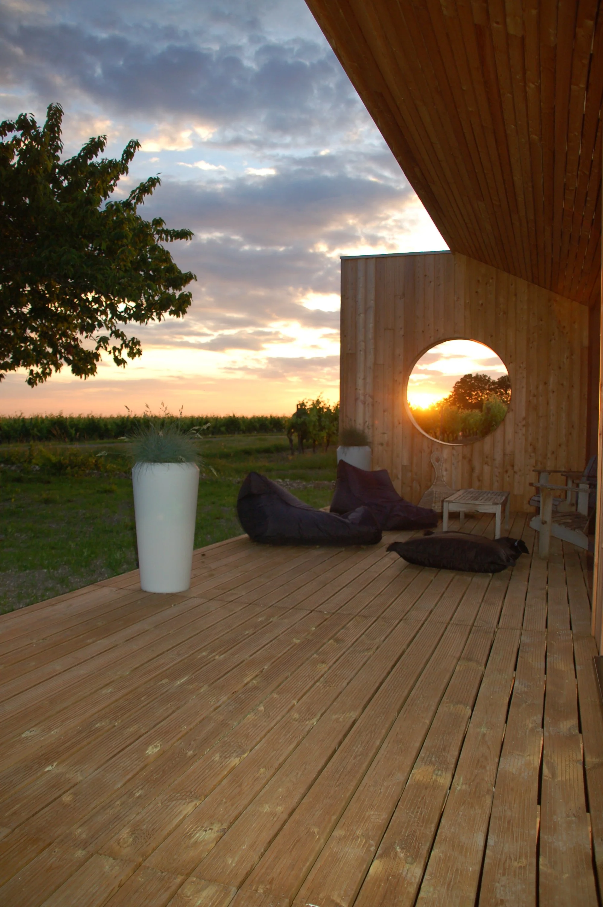 Vue d'une terrasse en bois avec un fauteuil, un coussin, une table en bois, et un mur en bois avec un trou circulaire, le tout au coucher du soleil avec le ciel partiellement nuageux.