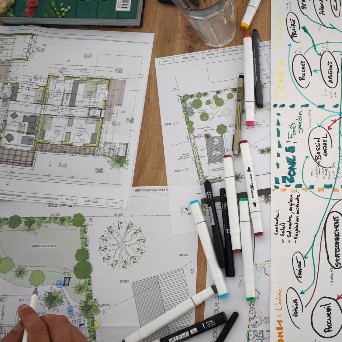 Plans architecturaux et esquisses pour un projet immobilier ou paysager, avec des marqueurs, une feuille blanche avec notes, et un verre d'eau sur une table en bois.