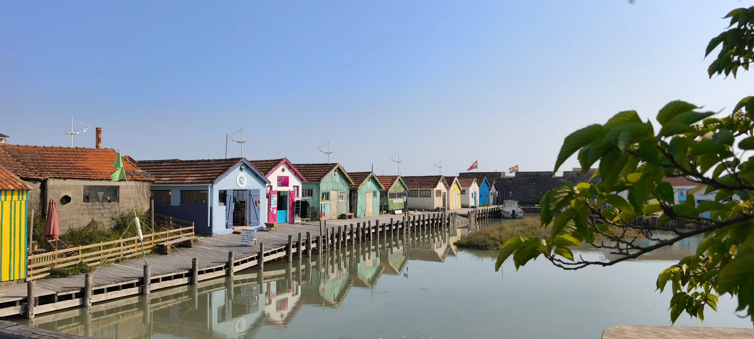 Maisons colorées en bois le long d'une jetée en bord de mer, avec des reflets dans l'eau calme, sous un ciel clair.