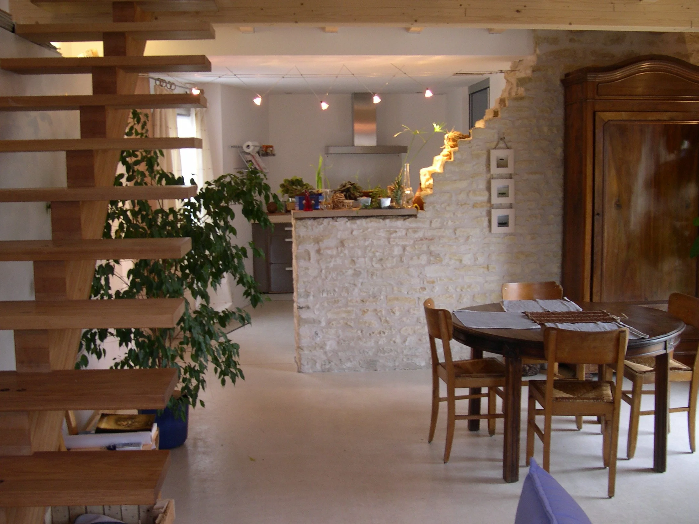 Intérieur d'une maison avec escalier en bois, table en bois avec chaises, mur en pierre, plantes vertes et lumière chaude.