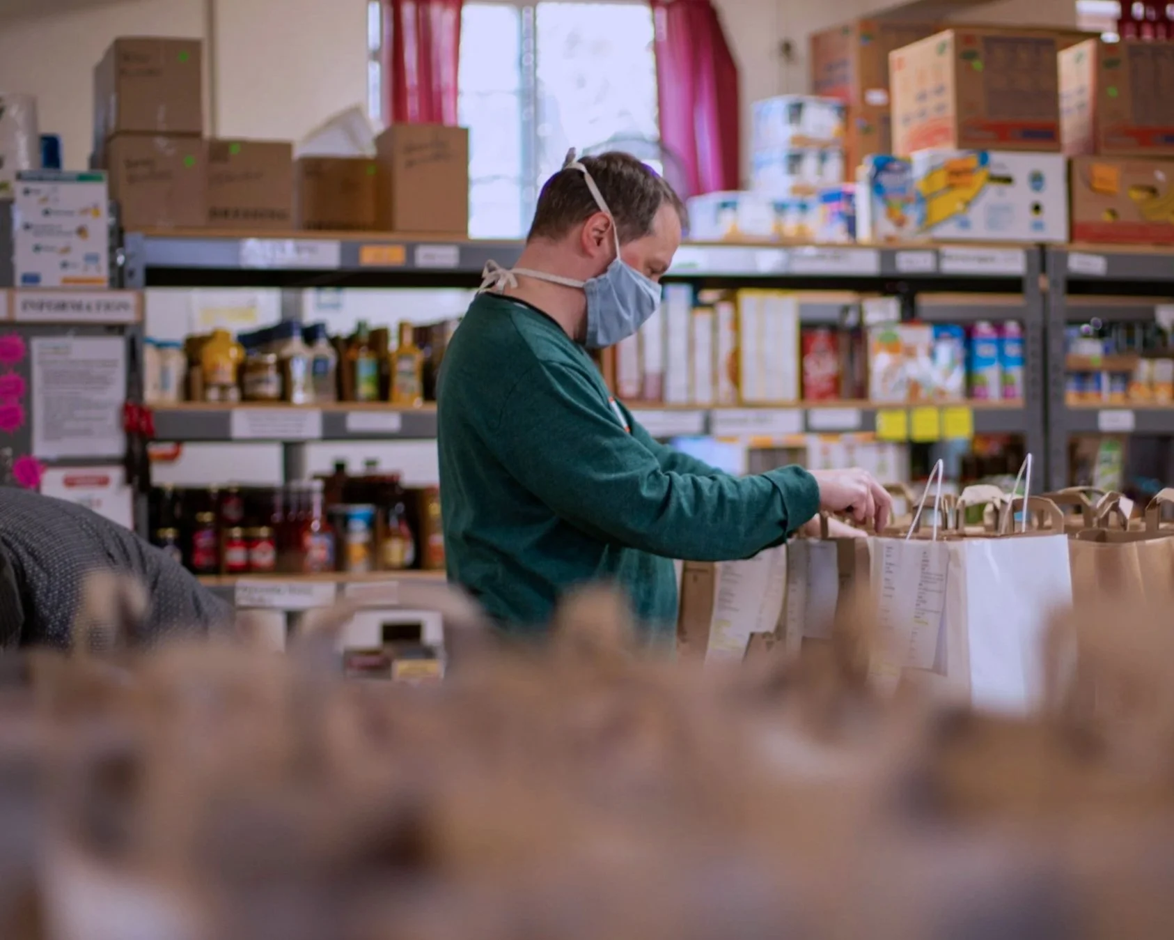 A person wearing a face mask and green sweatshirt shopping in a grocery store, reaching into paper bags. Shelves filled with various food and household items are visible in the background.