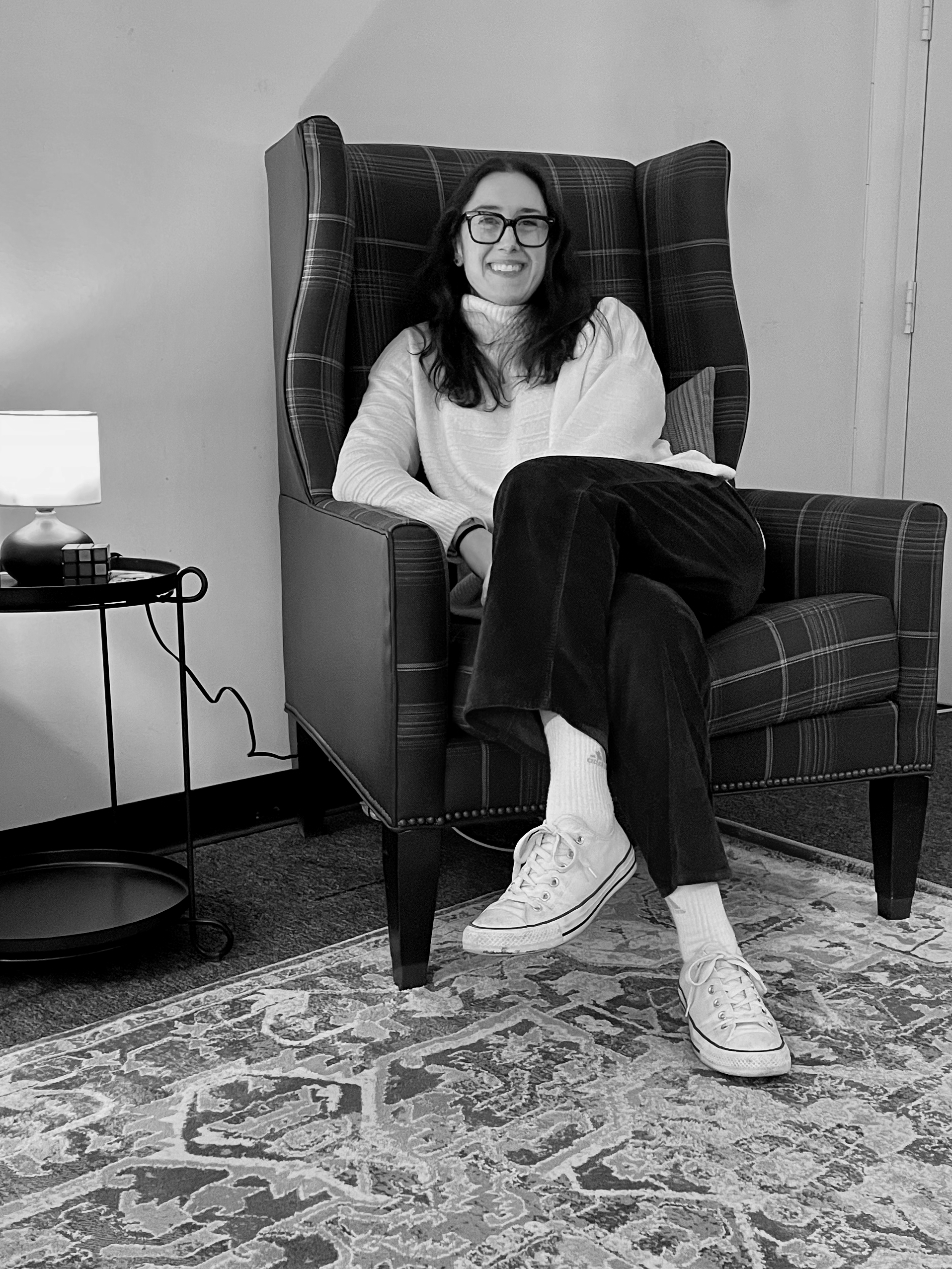 A woman with glasses and long dark hair sitting on a large, high-backed armchair, smiling. She is wearing a light-colored sweater, dark pants, white sneakers, and white socks, with a small pillow behind her. There is a side table with a lamp, a Rubik's cube, and other items beside her. The room has a patterned rug on the floor and a plain wall behind her.