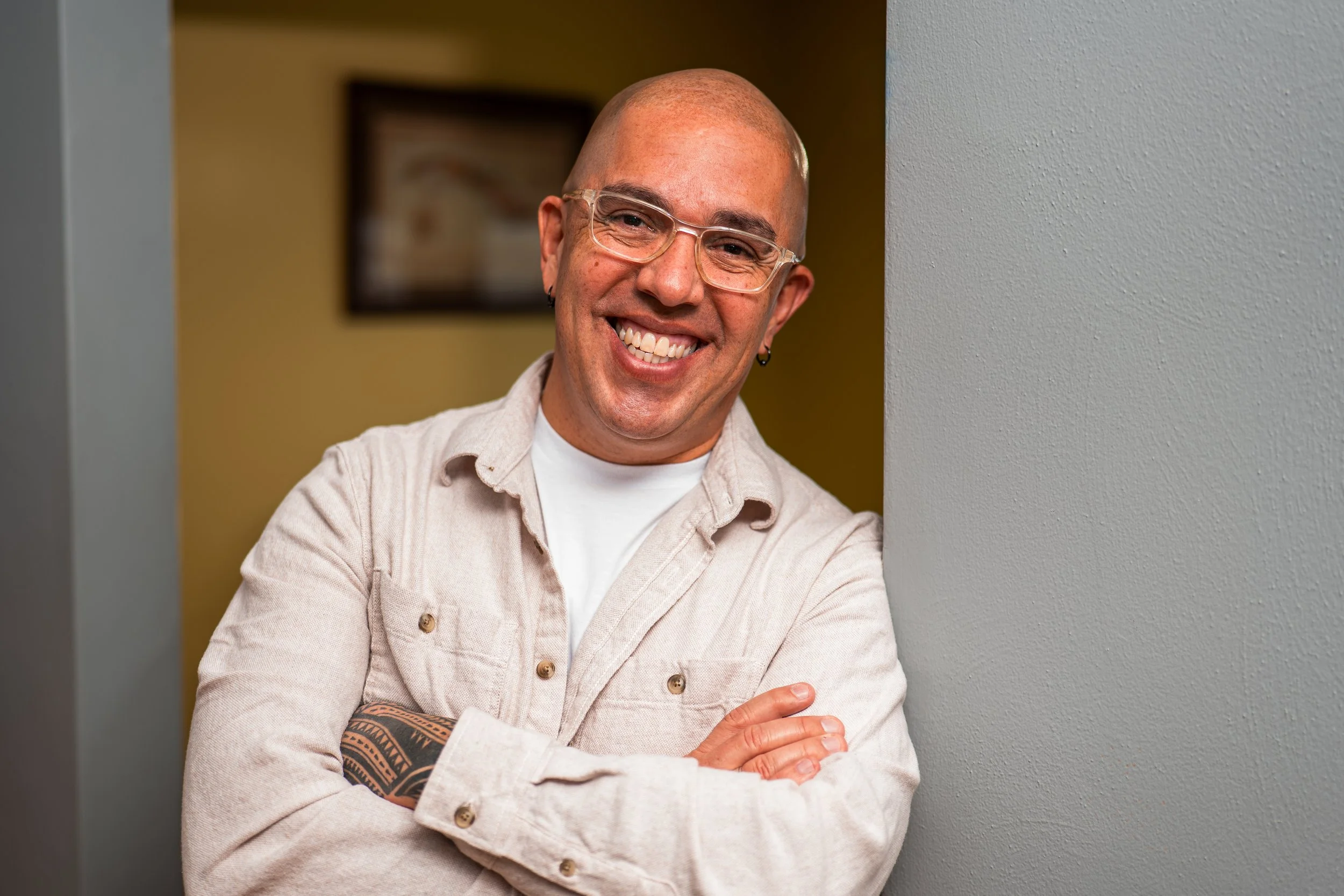 A smiling man with glasses and earrings, wearing a beige jacket with a tattoo on his arm, standing with arms crossed near a wall.