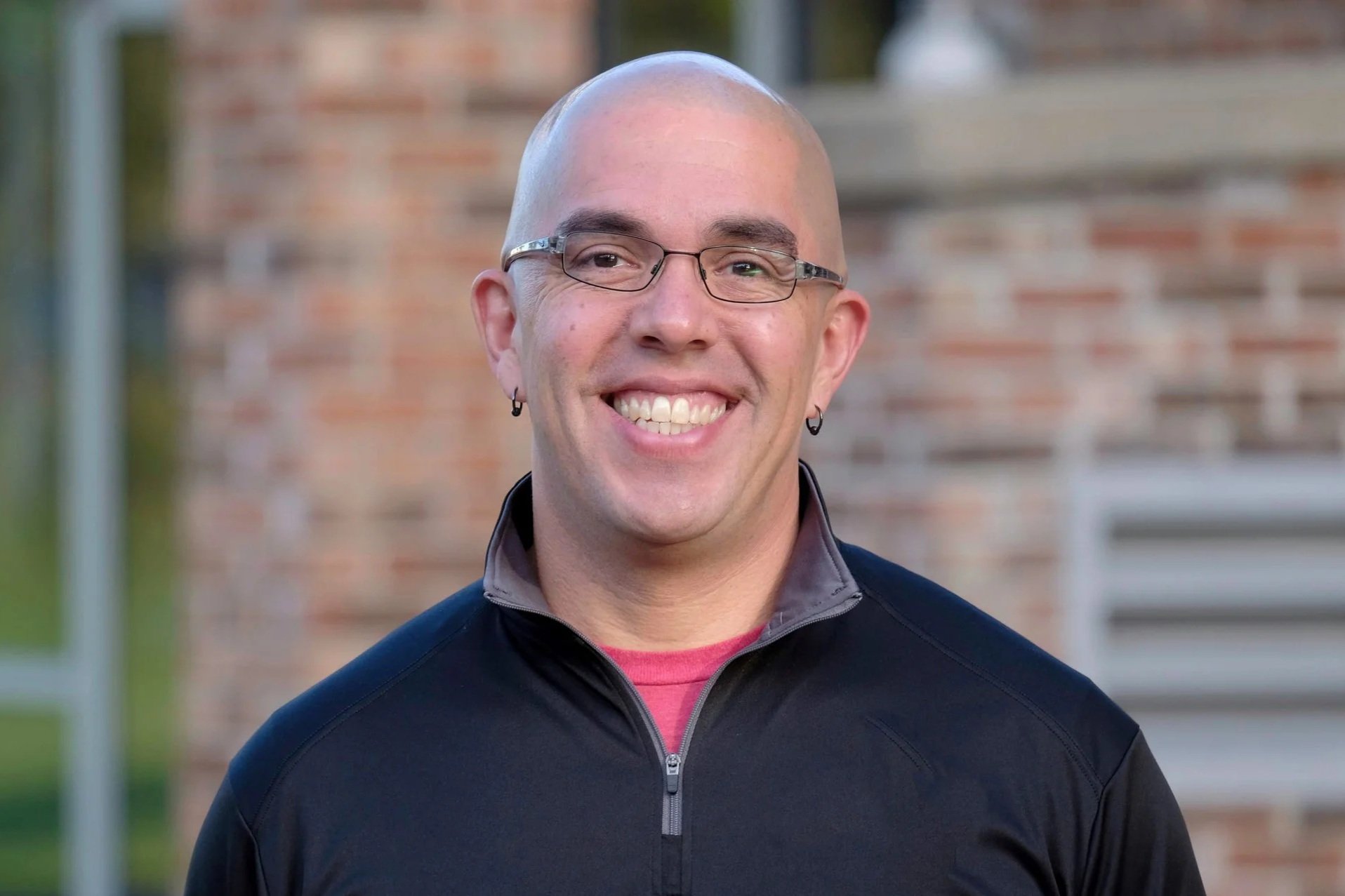 A smiling man with glasses, earrings, and a bald head standing outdoors in front of a brick wall.